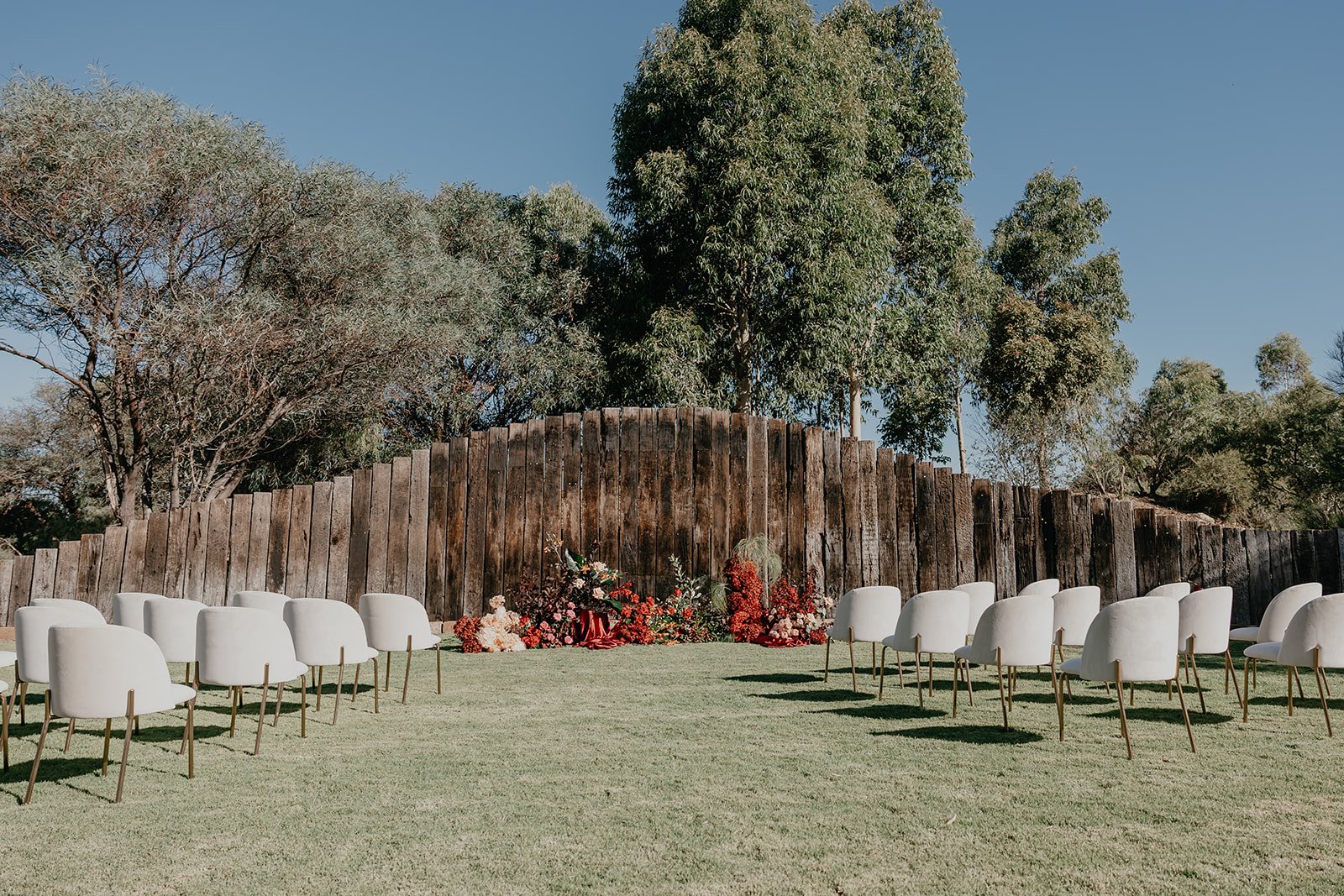 Outdoor wedding ceremony setup with white chairs on a grassy area, floral arrangements at the front, and a tall wooden fence with trees in the background on a clear day.