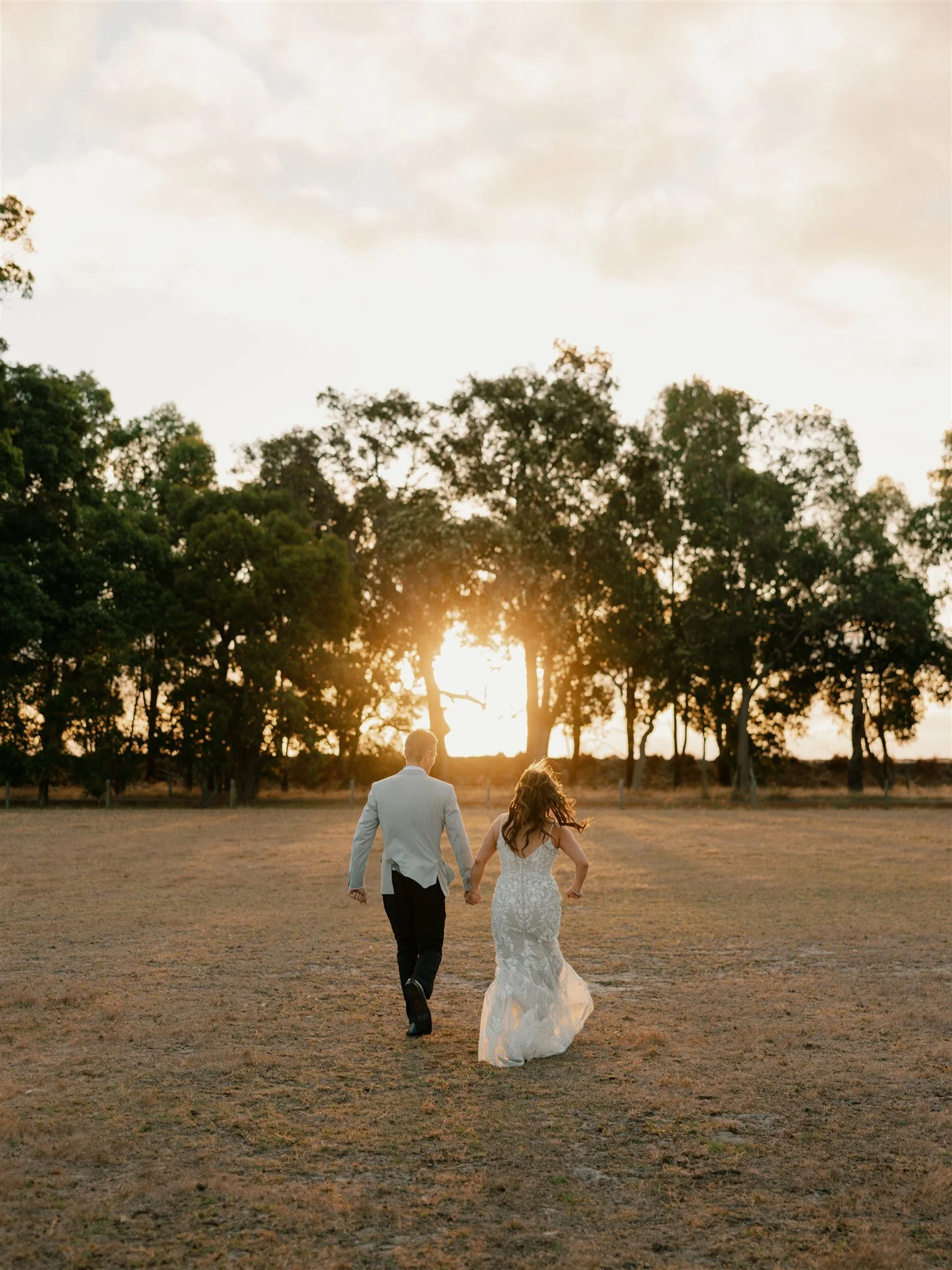 A couple in wedding attire running hand in hand across an open field at sunset with trees in the background.