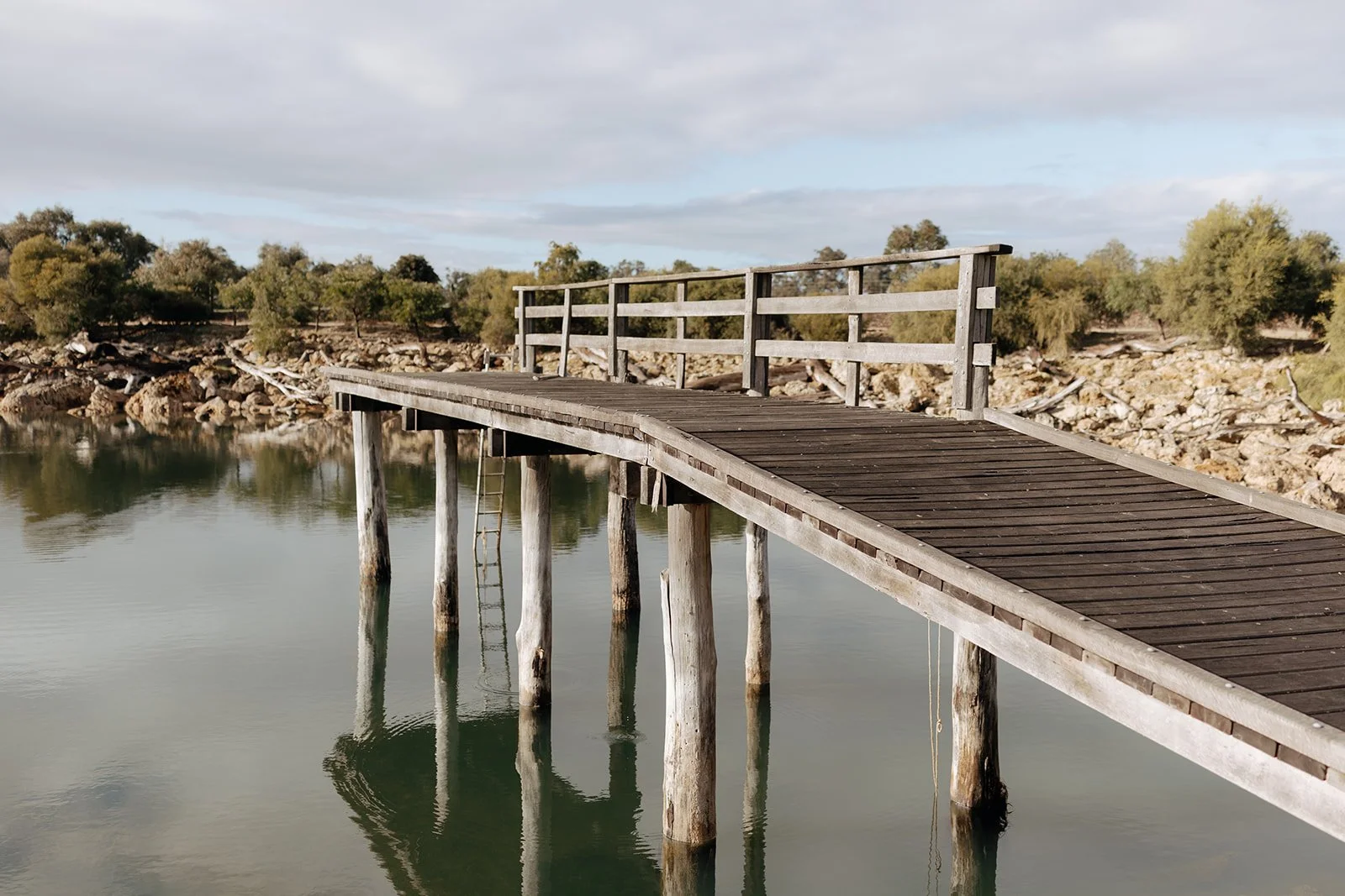 A wooden pier extending over calm water with a grassy, tree-lined shoreline in the background.