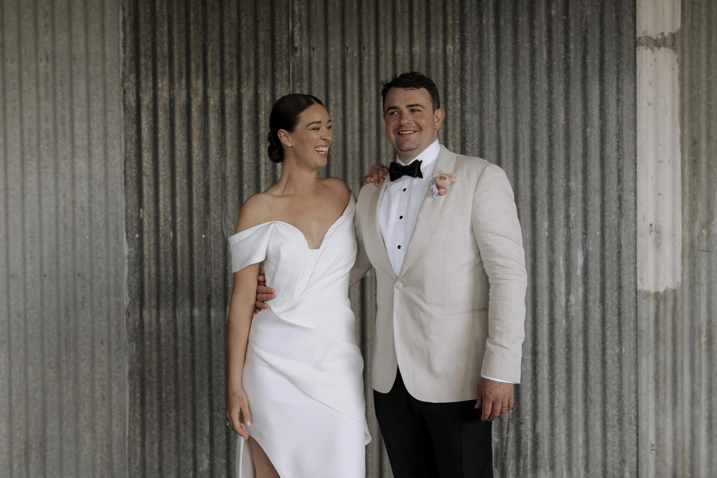 A bride in a white wedding dress and a groom in a beige tuxedo with a black bowtie standing together indoors, smiling and looking at each other against a corrugated metal wall background.