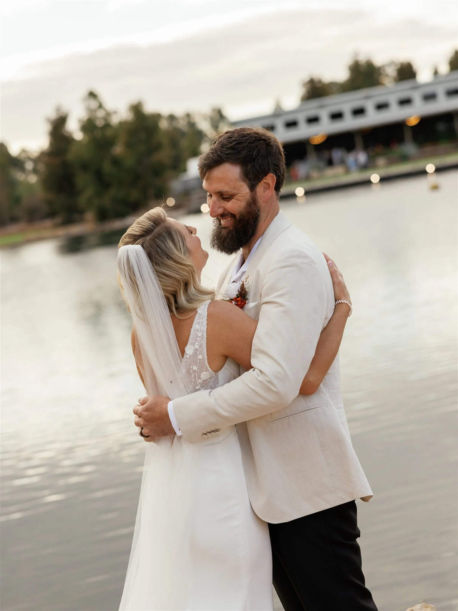 A newlywed couple embracing outdoors near a body of water, with a bridge and trees in the background during sunset.