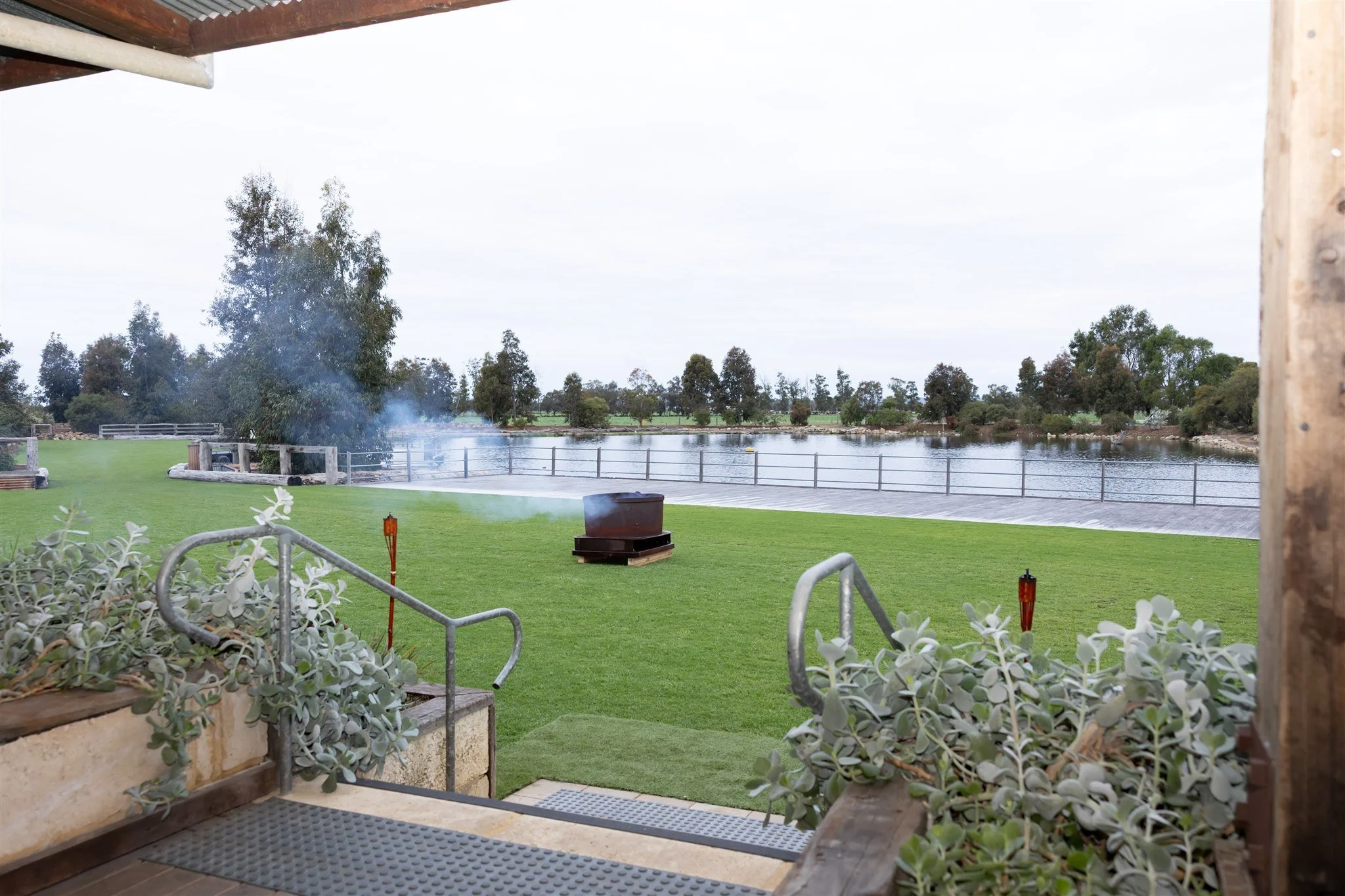 View from a patio overlooking a grassy area by a body of water, with a fire pit emitting smoke, trees in the distance, and some decorative plants in the foreground.