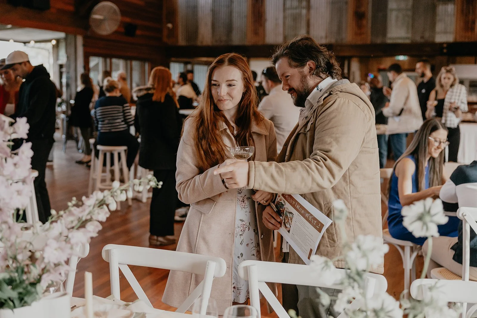 A woman with long red hair and a man with wavy brown hair and beard are talking at a social event, holding a drink and a menu. The background shows other people mingling and sitting at tables in a rustic indoor setting with wooden walls.