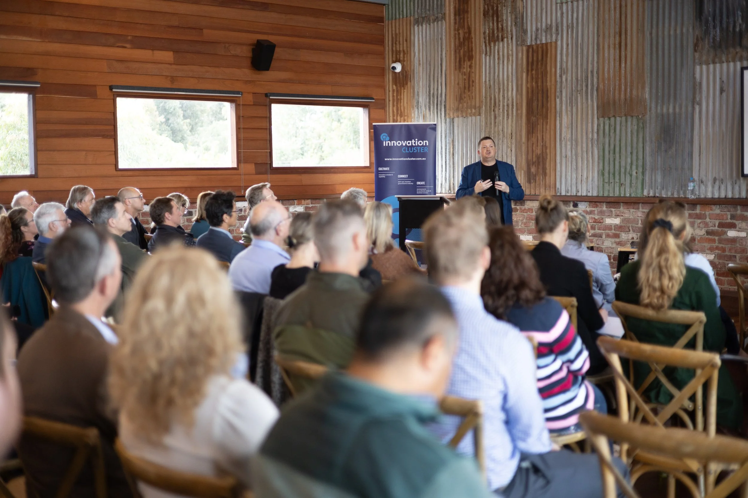 A man in a blue blazer giving a presentation to an audience seated in a room with wooden and brick walls, windows, and a blue banner that reads 'Innovation Cluster'.