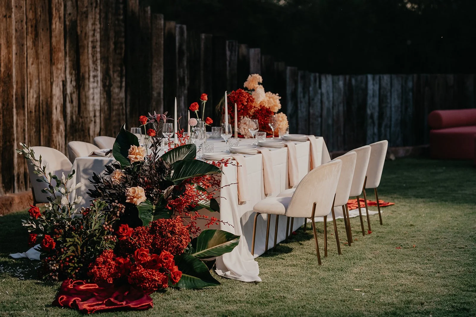 An outdoor table decorated for a special event with floral arrangements and tableware, set against a wooden fence with a grassy ground.