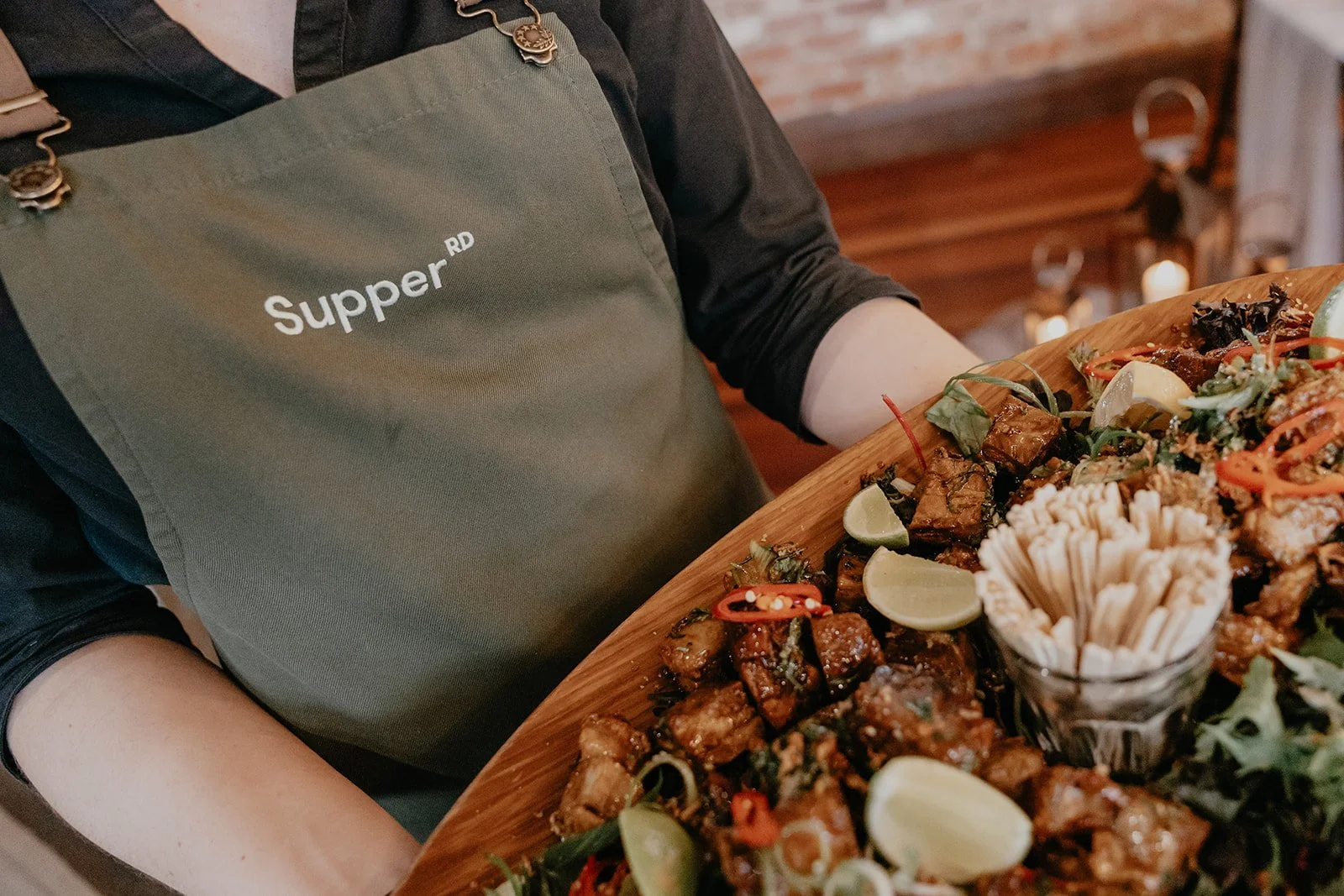A person wearing a green apron with the word 'Supper RD' and a black shirt is holding a large wooden tray filled with various Asian dishes, including pieces of meat, lime wedges, and a jar of aspen sticks, in a restaurant setting with warm lighting and a brick wall background.