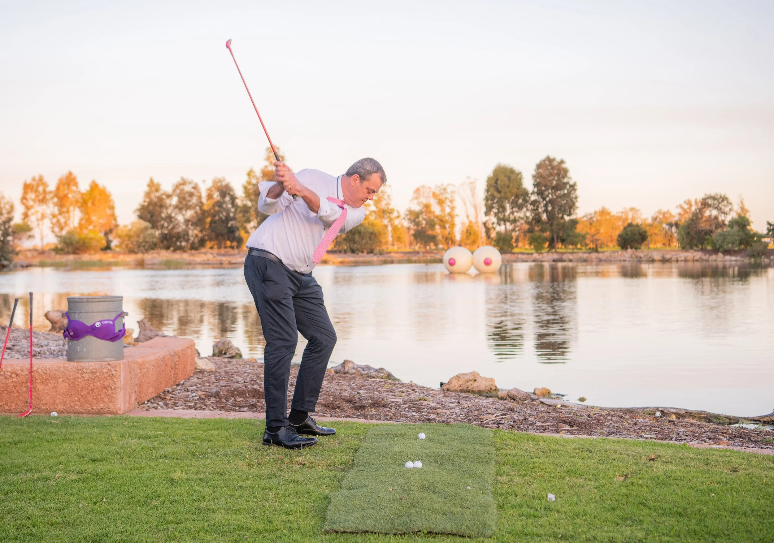 A man in business attire preparing to hit a golf ball on a golf course near a body of water during sunset.