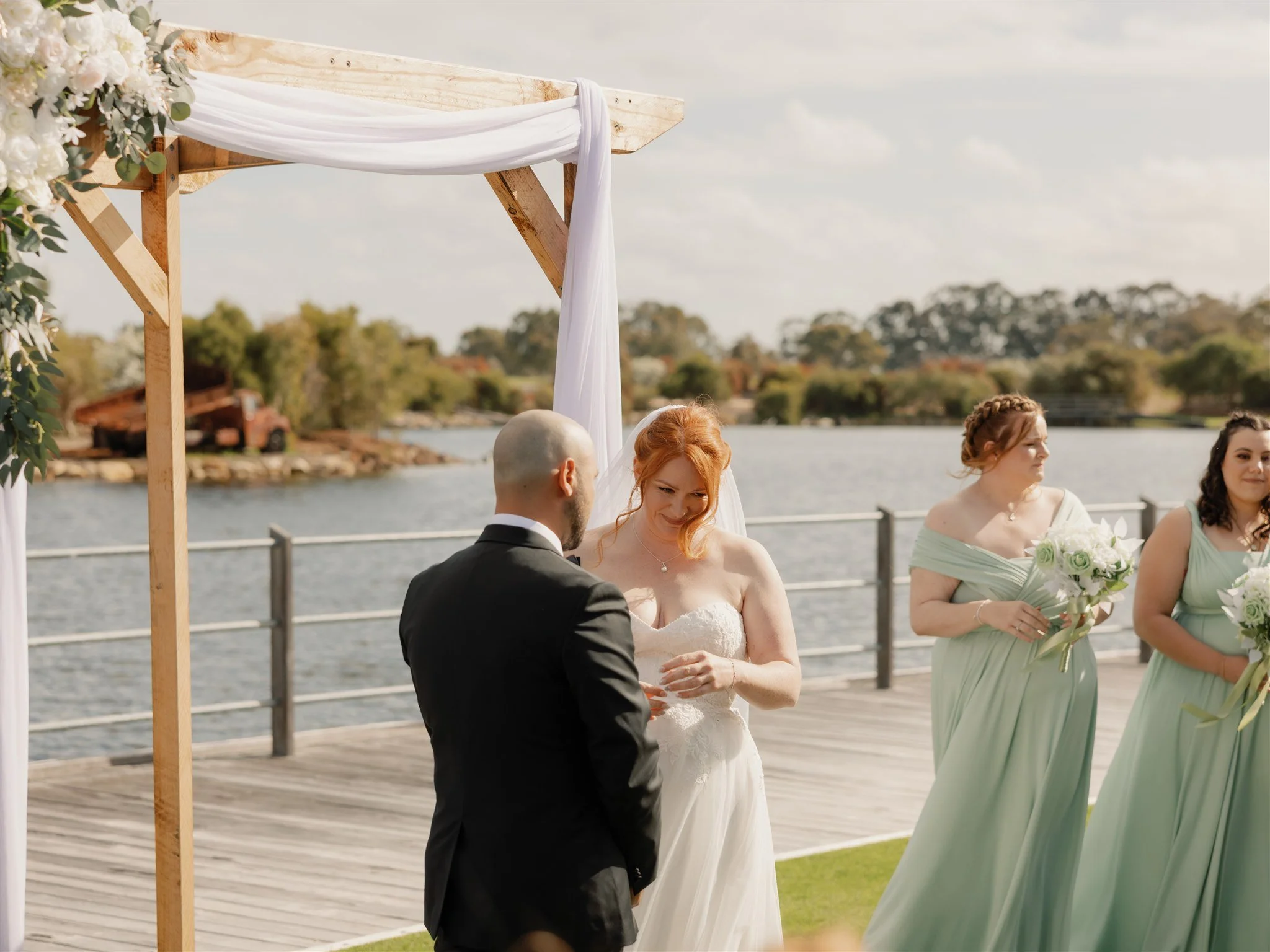 A wedding ceremony taking place outdoors by a river with a wooden arch decorated with white fabric, with the bride and groom exchanging vows, and bridesmaids holding bouquets in the background.