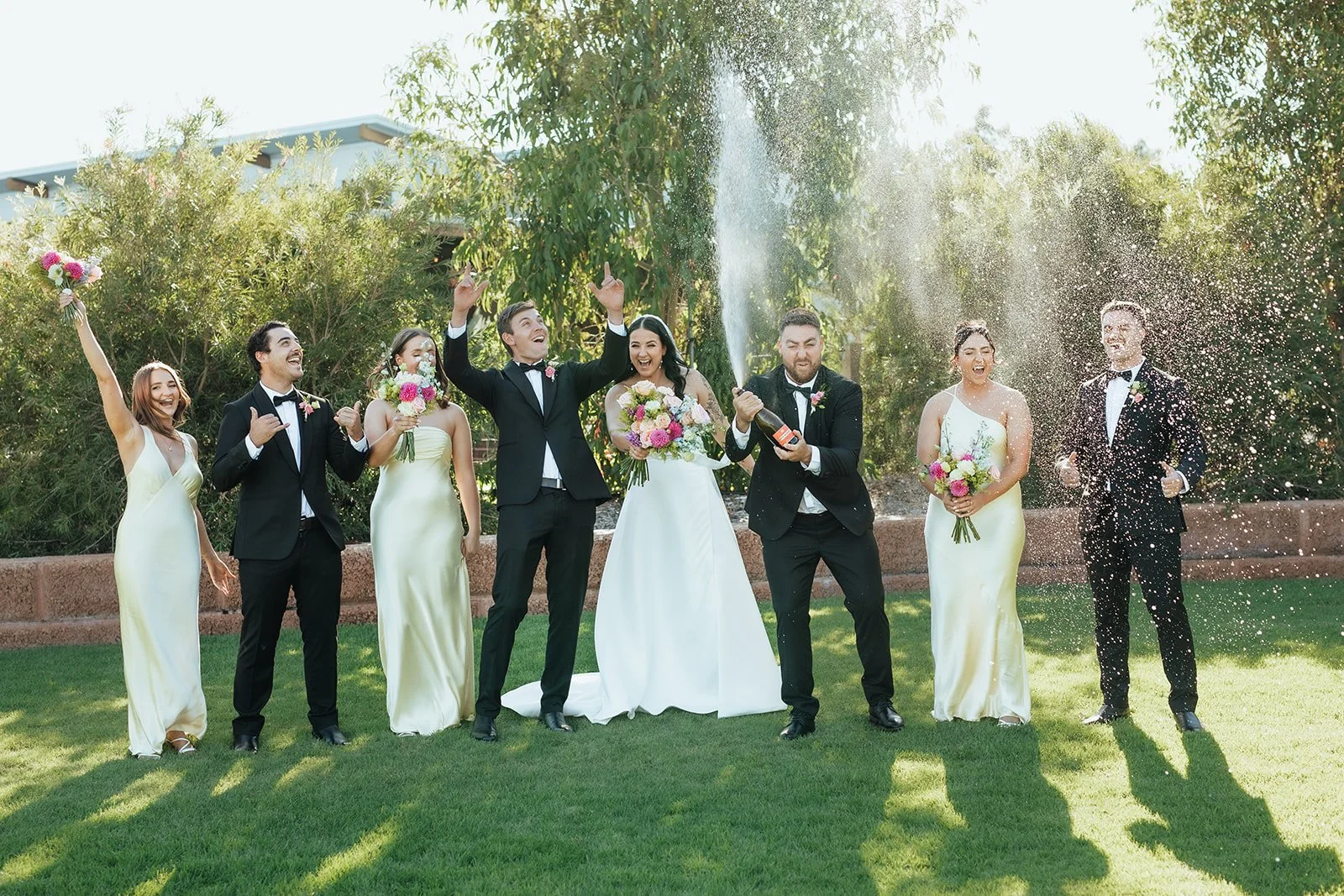 A wedding celebration outdoors with a bride, groom, bridesmaids, and groomsmen. The bride and groom are in the center, with the groom opening a bottle of champagne. Groomsmen are wearing black tuxedos, and bridesmaids are in light yellow dresses. They are all smiling and celebrating on a sunny day, with confetti or spray from the champagne in the air.