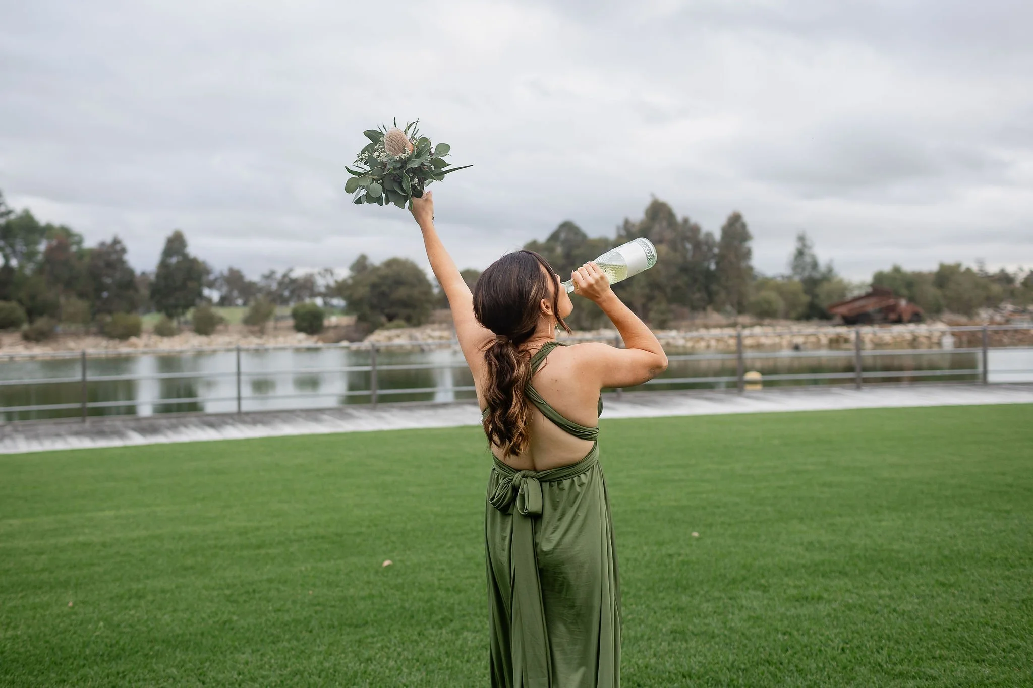 A woman in a green dress holding a bouquet of flowers in one hand and a bottle in the other, standing on a grassy outdoor area near a body of water, with trees and a cloudy sky in the background.
