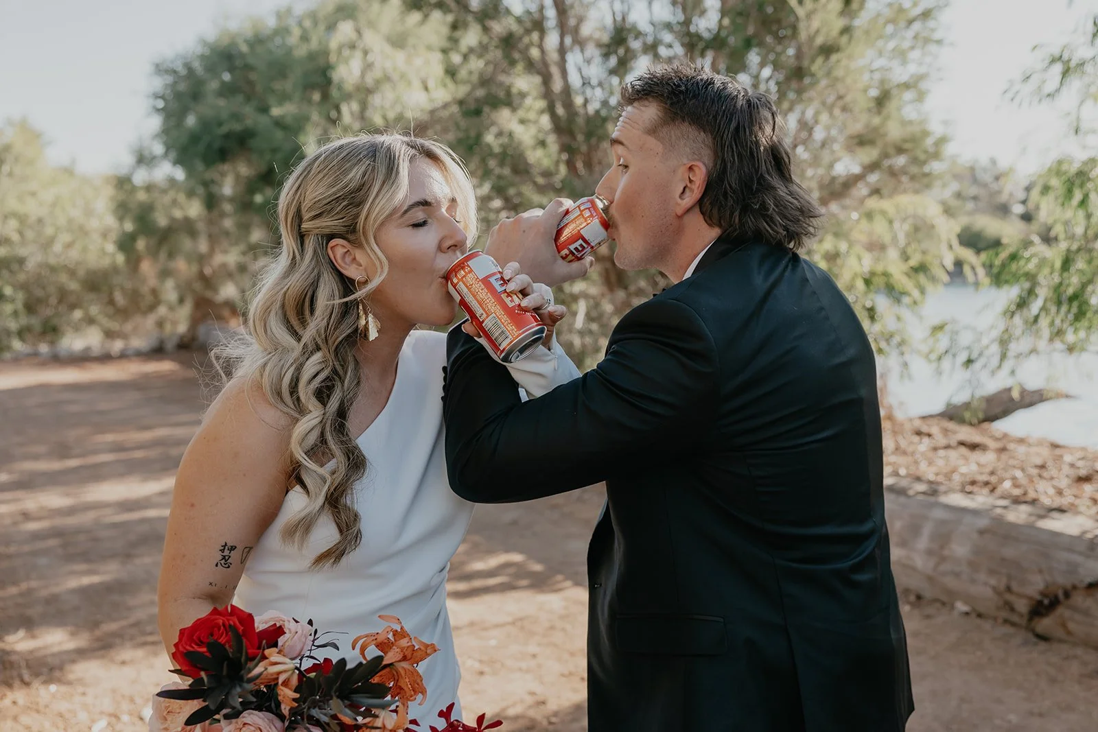 A newlywed couple sharing beers outdoors, the bride holding a bouquet of flowers, in a natural setting near a body of water.