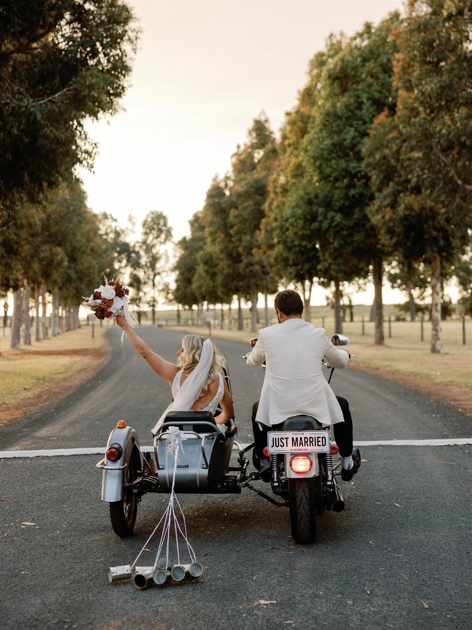 A newly married couple riding a motorcycle with a sidecar, the bride holding a bouquet and wearing a wedding dress and veil, the groom wearing a white suit, on a rural road surrounded by trees at sunset, with a 'Just Married' sign on the back of the motorcycle and cans tied to the sidecar.