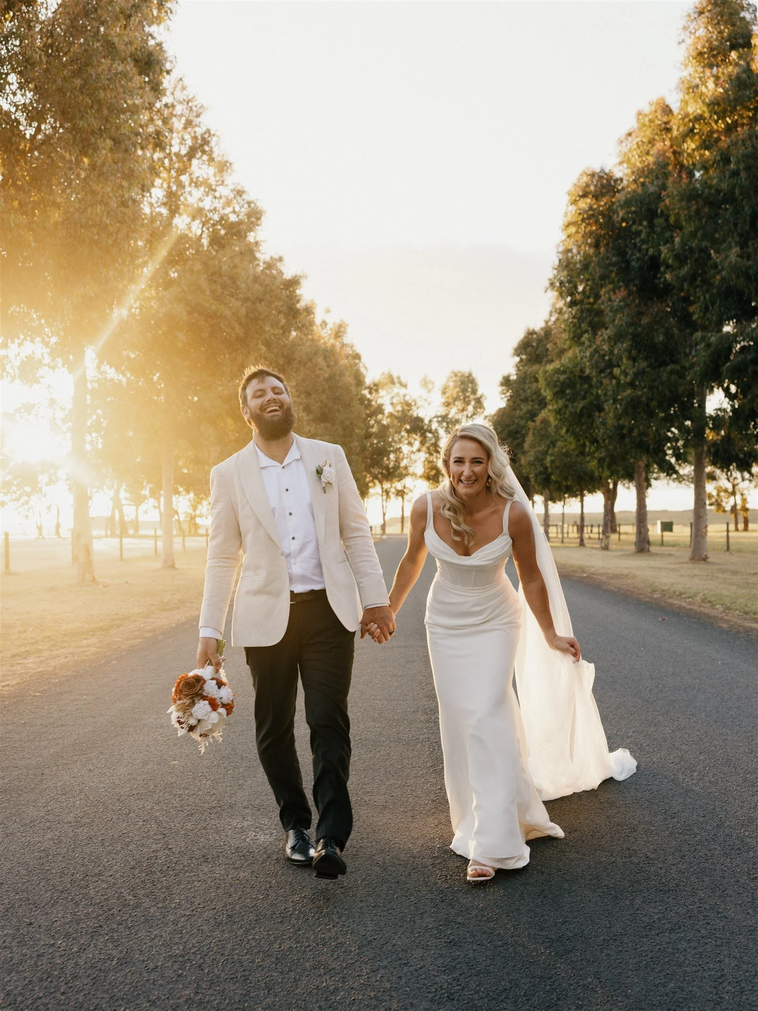 A newlywed couple holding hands and walking down a road at sunset, with trees on each side. The groom is dressed in a cream-colored suit jacket, white shirt, and black pants, and is holding a bouquet. The bride is in a white wedding dress with a train and is smiling while holding up her dress slightly.