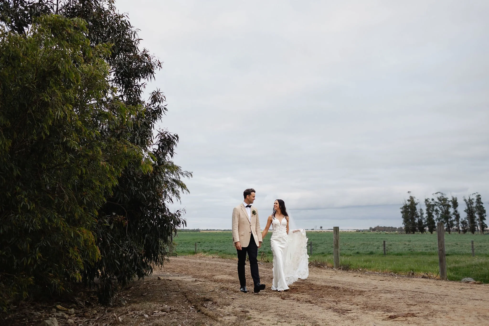 A bride and groom walking hand in hand on a rural dirt path, with green fields and trees in the background under a cloudy sky.