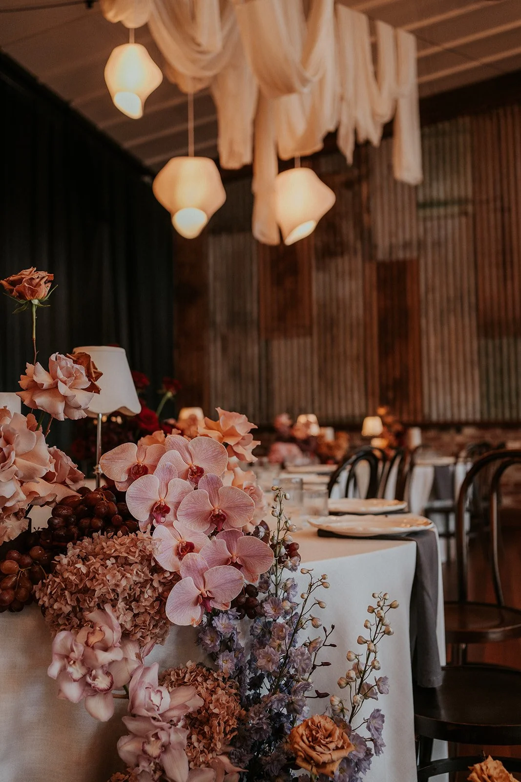 Elegant banquet table decorated with pink and purple flowers, set in a warmly lit room with wooden walls and ceiling, and modern hanging light fixtures.