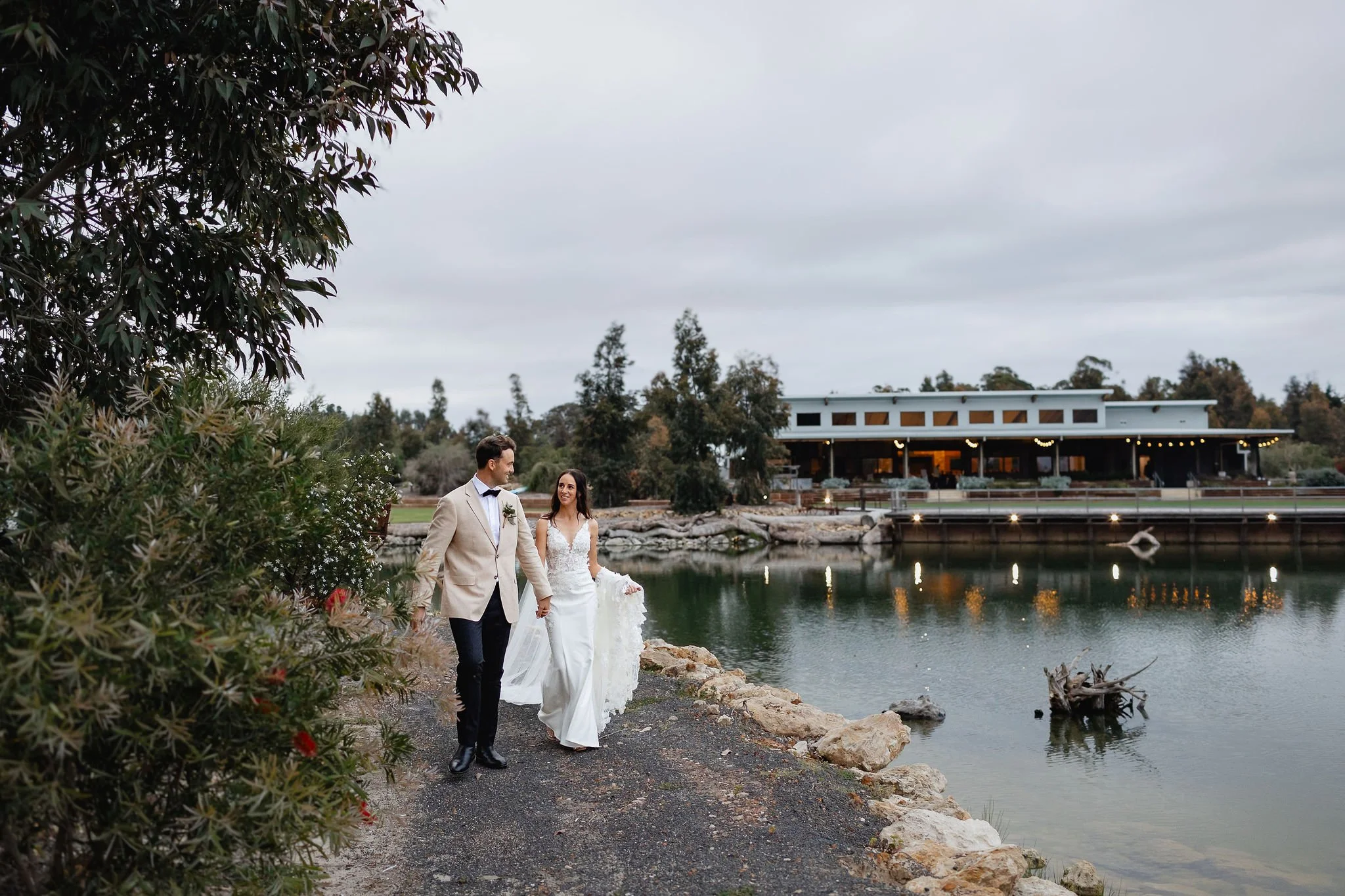 A newlywed couple walking hand in hand along a lakeside path, with a modern building and trees in the background on a cloudy day.