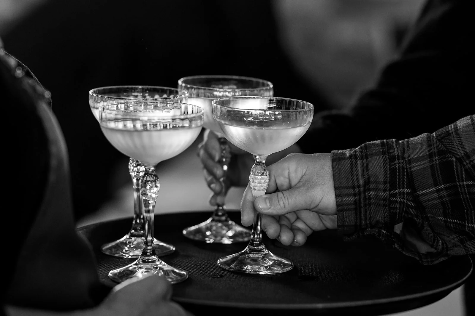 A black and white photo of several glasses of champagne being held by people. One person wears a plaid shirt.