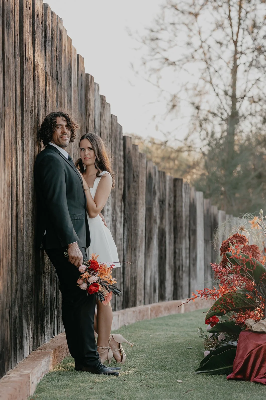 A man and woman standing close together outdoors near a wooden fence, with trees in the background. The man is holding a bouquet of flowers, and the woman is resting her hand on his chest, wearing a white dress, while he is dressed in a black suit.
