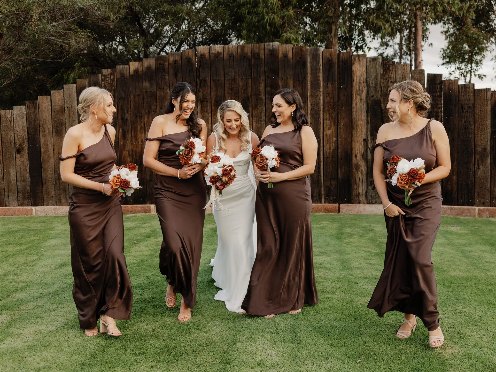 Wedding bride and bridesmaids standing on grass with brown wooden fence in background, holding bouquets.