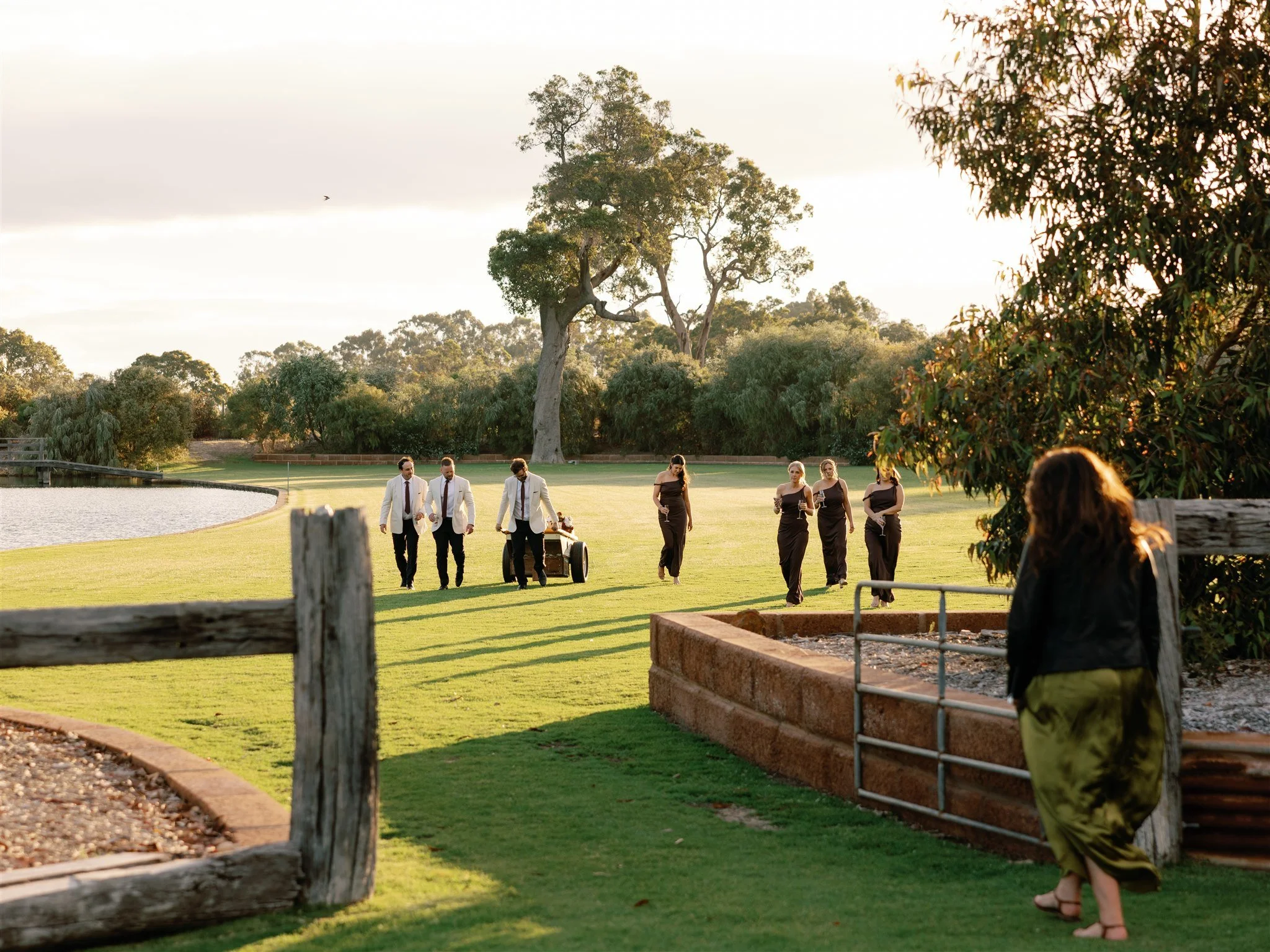 A group of people dressed in formal attire walking on a grassy area near a lake, with trees in the background and a person standing near a garden bed in the foreground.