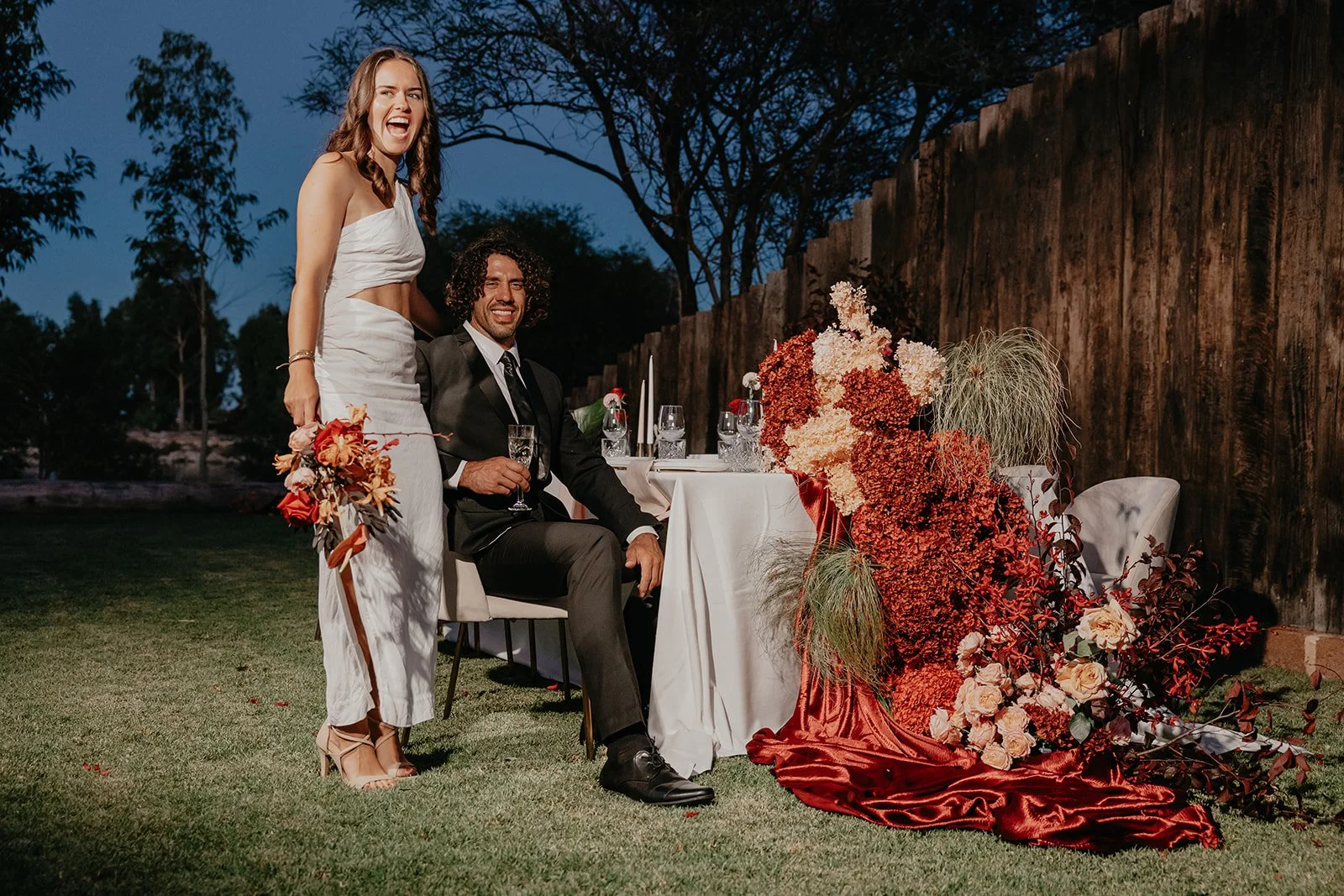 A woman in a white dress holding a bouquet and a man in a suit sitting at a decorated outdoor table during dusk with floral arrangements and candles.