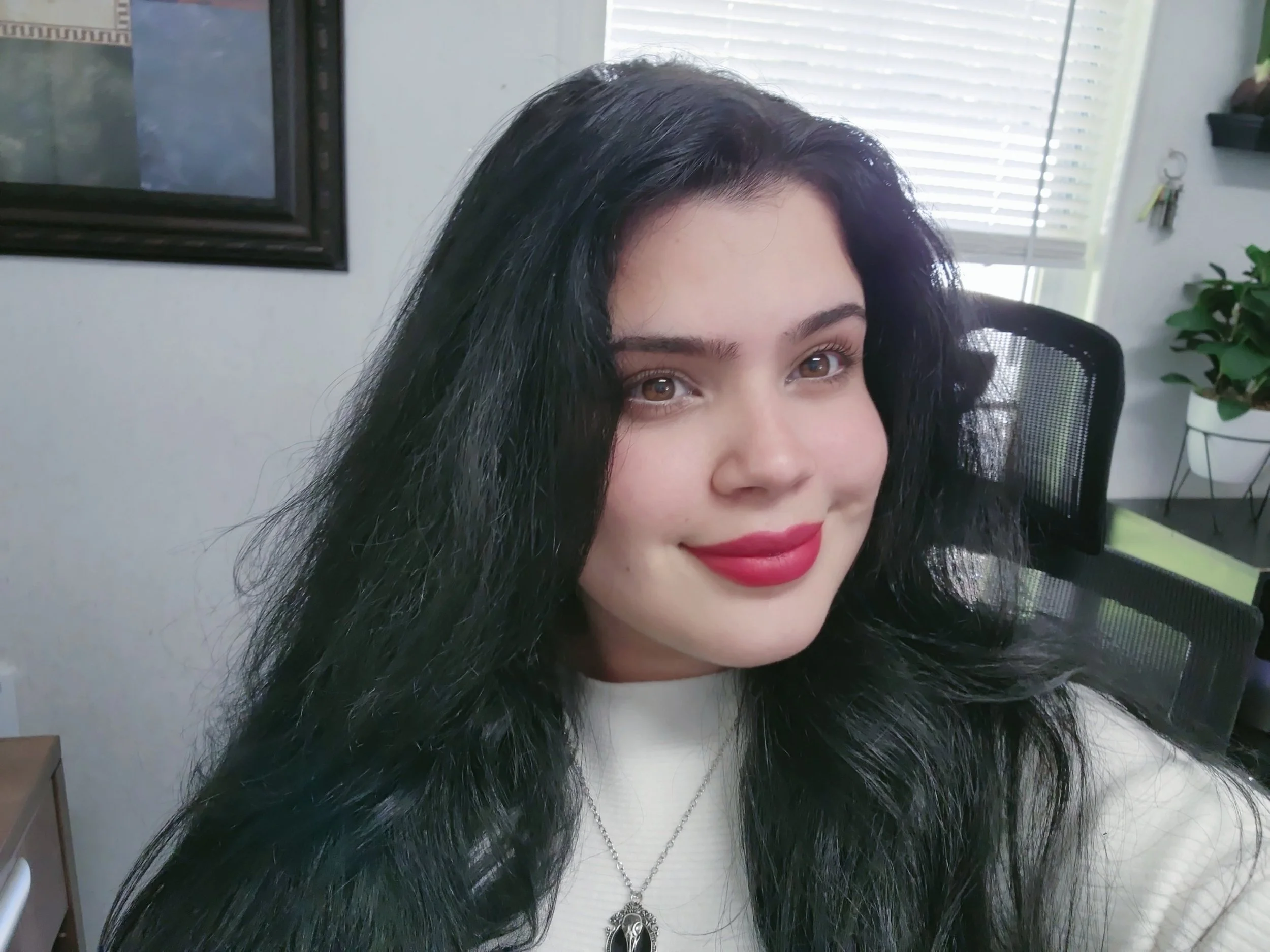 A woman with long black hair and red lipstick smiling at the camera in an office setting.