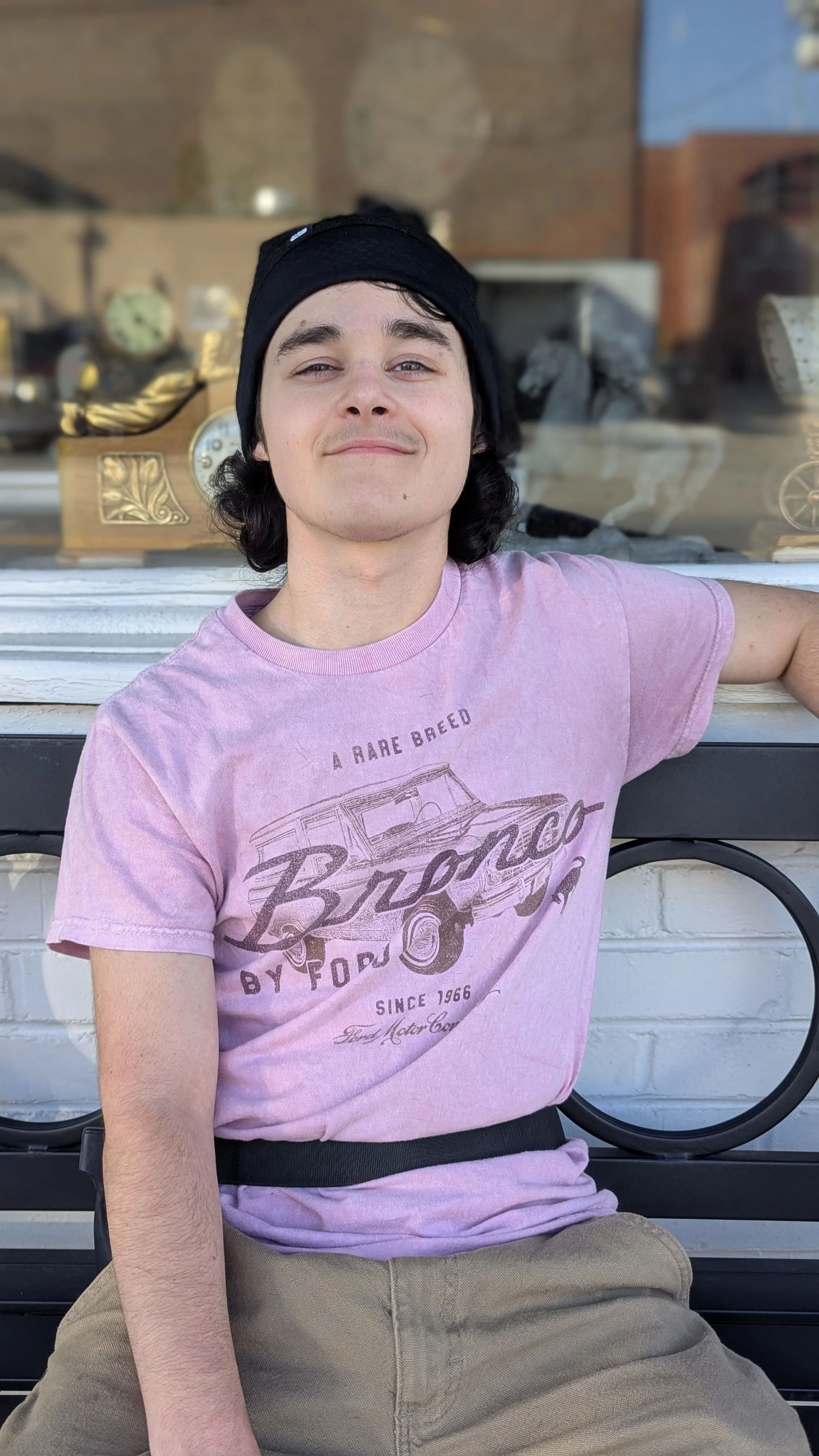 A young man with black hair, wearing a black beanie and pink t-shirt, sitting on a bench outside, smiling.