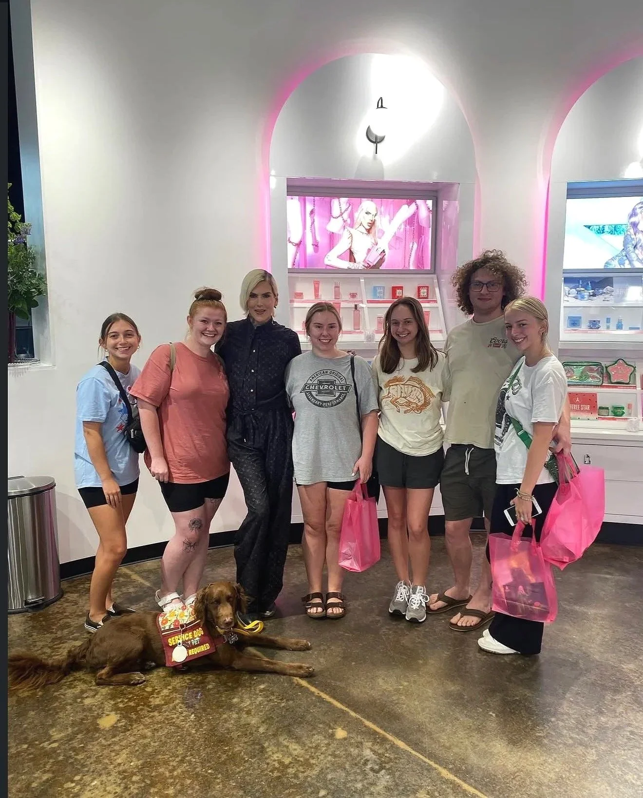 Service Dog "Oso" relaxing on the floor, in a down stay amongst a group of people standing inside a retail store with pink lighting accents, shelves with products, and digital screens in the background - Celebrating a Jeffrey Star makeup launch.