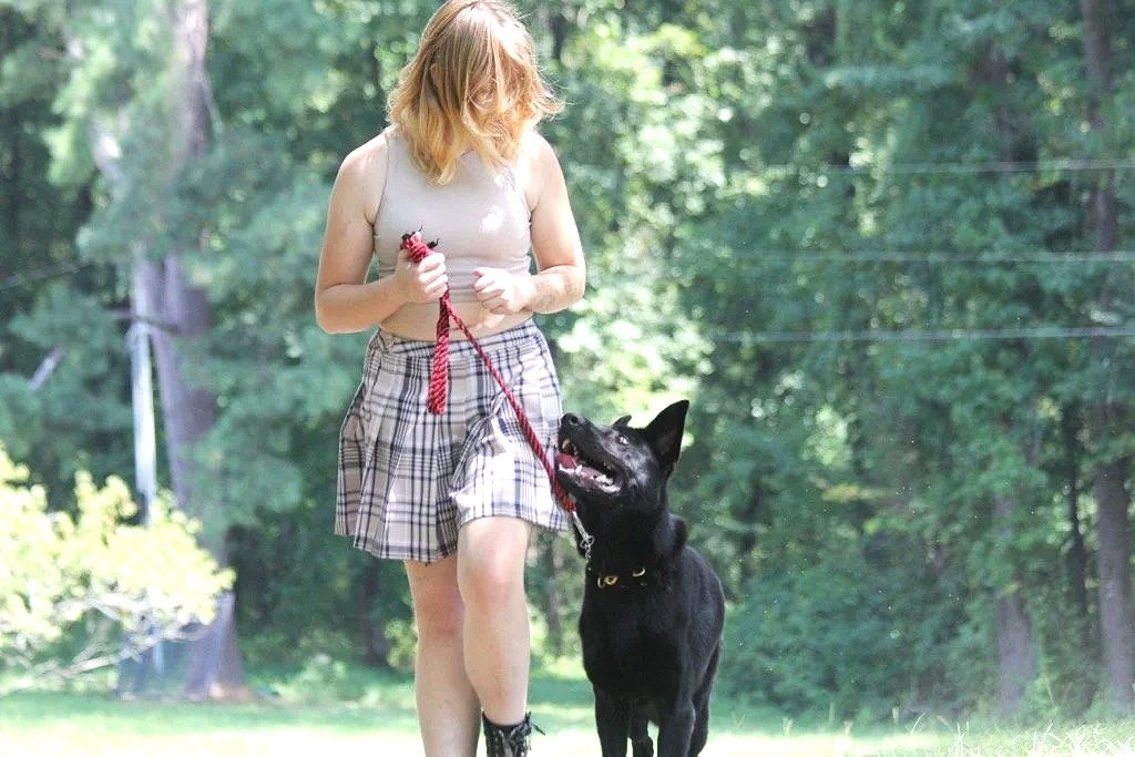 A woman with red hair walking a black dog on a leash outdoors in a green park or wooded area.