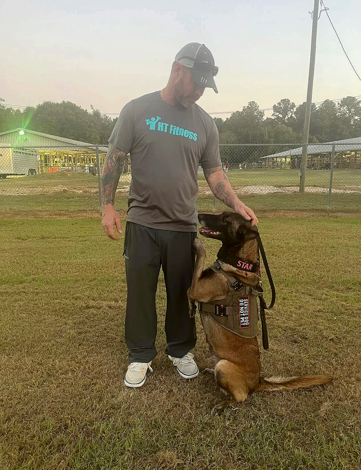 A man in a gray T-shirt with 'HT Fitness' printed on it, wearing a cap, pants, and sneakers, standing on grass. He is petting a Belgian Malinois police dog that is wearing a vest labeled 'SERVICE DOG' and 'DO NOT PET,' sitting with one paw on the man's hand and looking up at him. There are trees, a chain-link fence, and a building in the background.