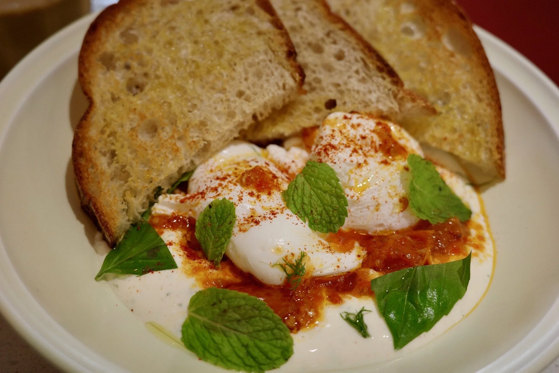 Poached eggs topped with red pepper flakes and fresh mint leaves, served with toasted bread slices on a white plate.