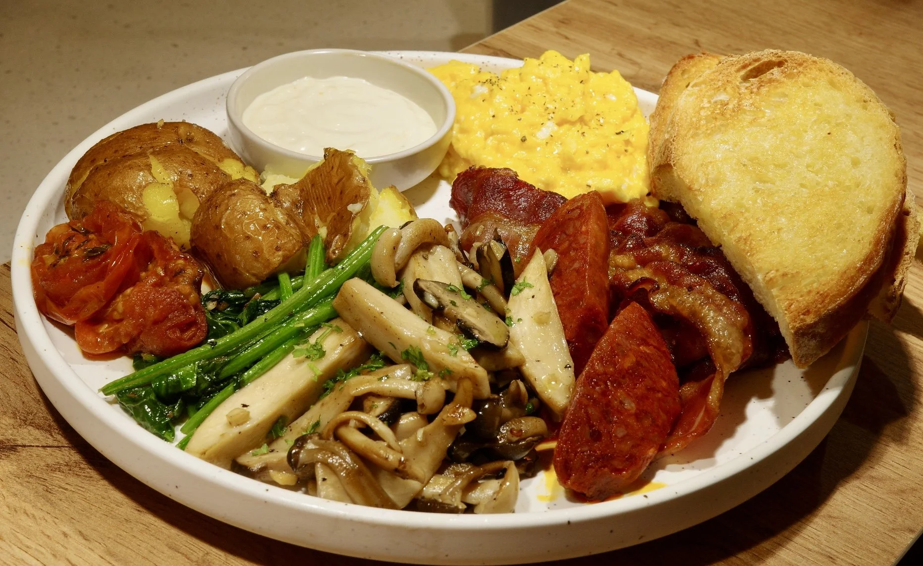 A breakfast plate with scrambled eggs, bacon, toast, roasted potatoes, sautéed mushrooms and greens, and a small bowl of yogurt or sour cream