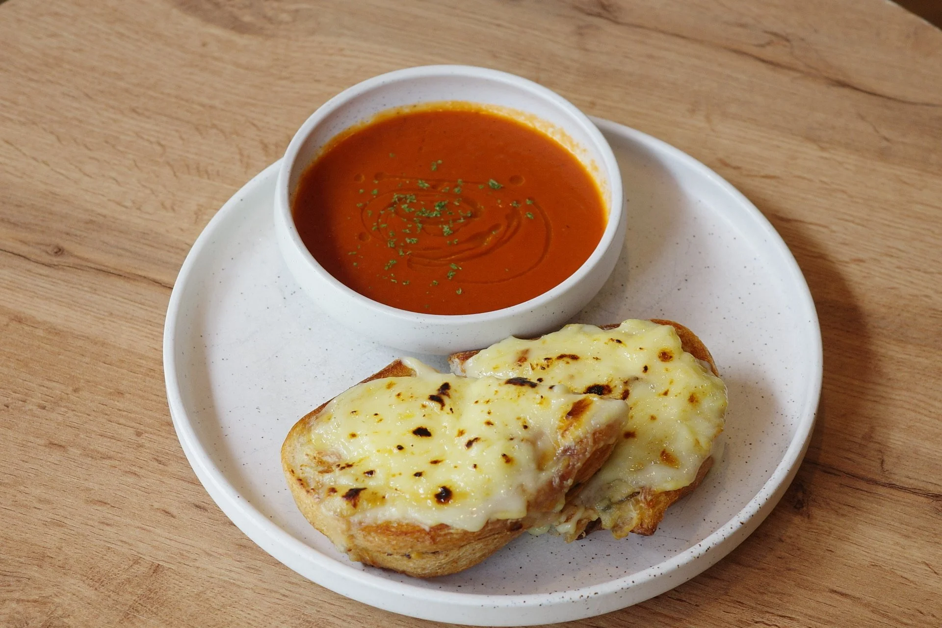 A white plate holds a slice of garlic bread with melted cheese, served with a cup of tomato soup garnished with herbs, on a wooden table.