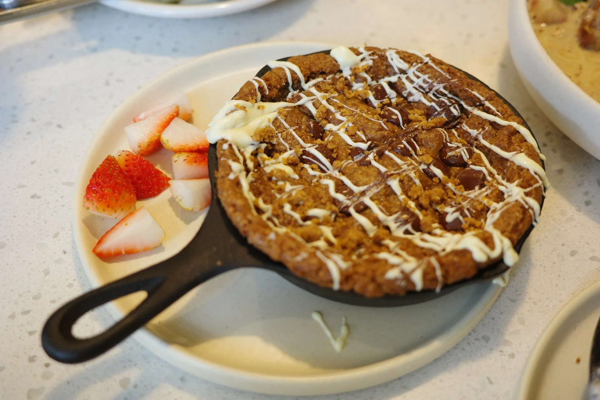 A chocolate chip skillet cookie topped with white chocolate drizzle, served with strawberries and strawberries on the side.