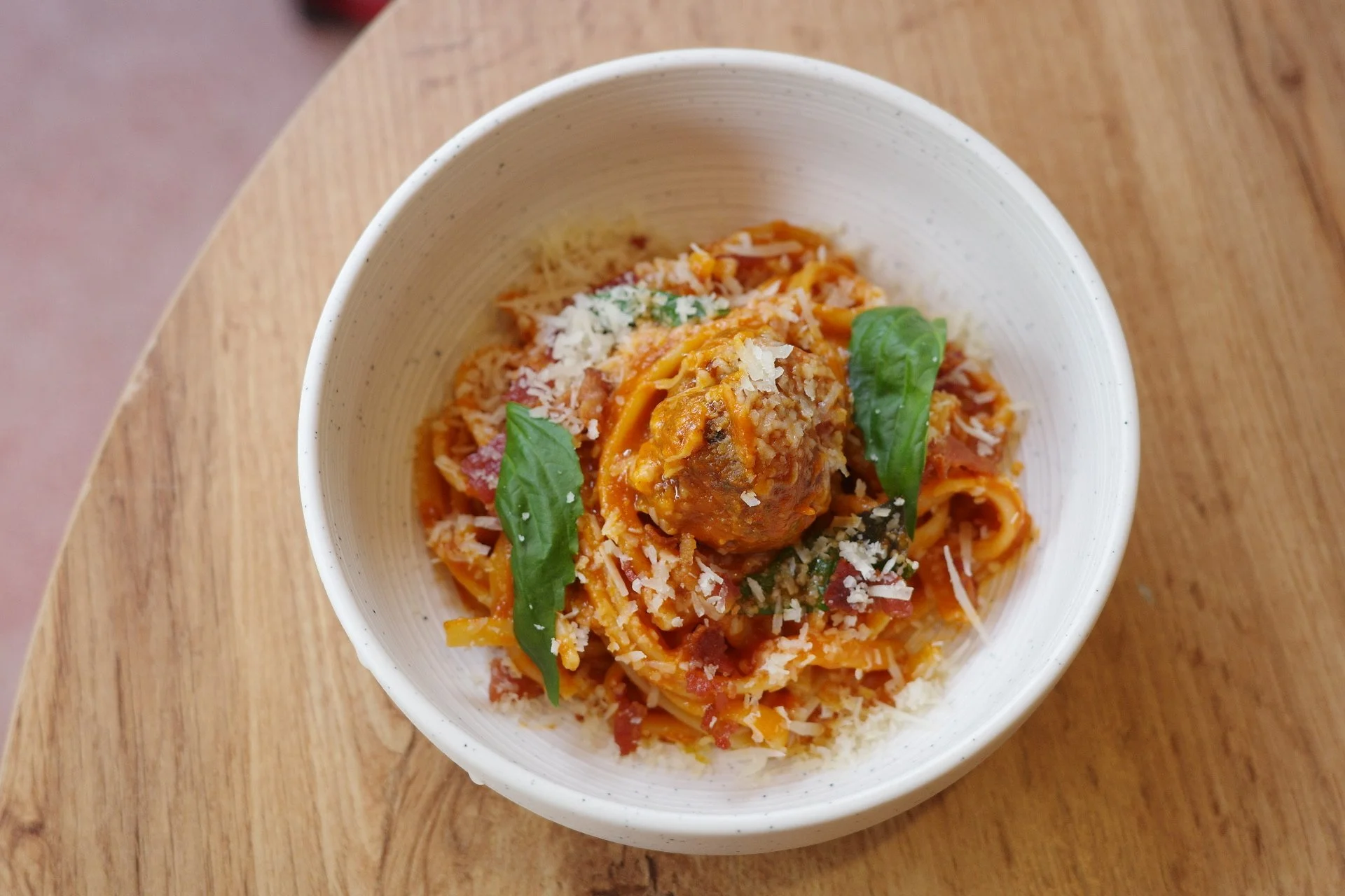 A bowl of spaghetti with meatball, topped with grated cheese and fresh basil leaves, on a wooden table.