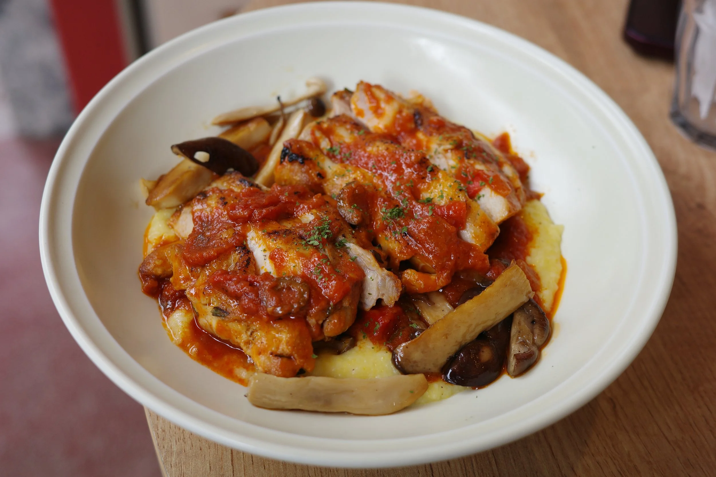A bowl of mashed potatoes topped with cooked chicken breast in red marinara sauce, garnished with herbs, and accompanied by sautéed mushrooms.