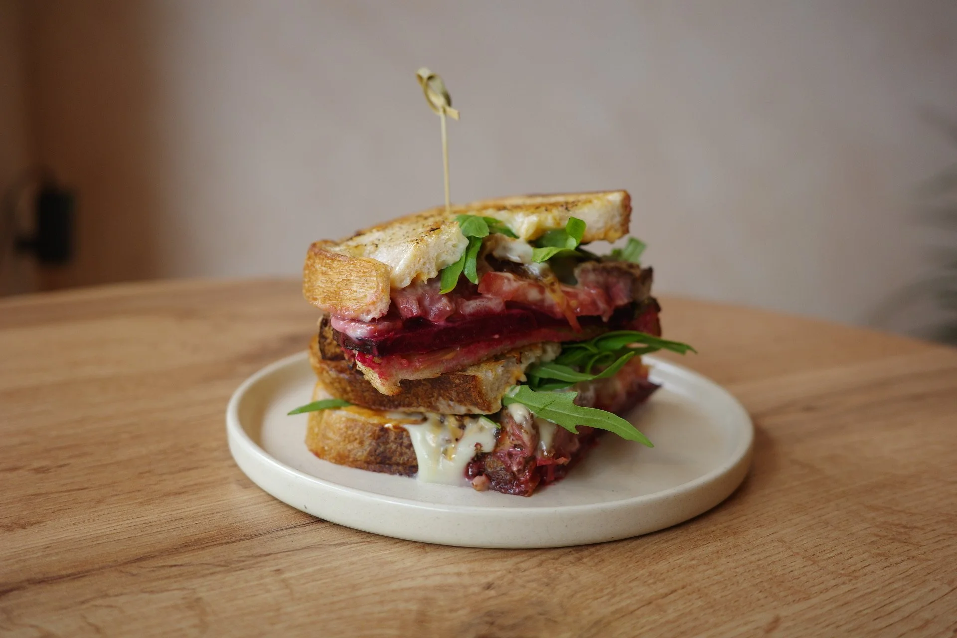 Stacked sandwich with various meats, cheese, lettuce, and a fried egg on top, served on a small white plate over a wooden surface.