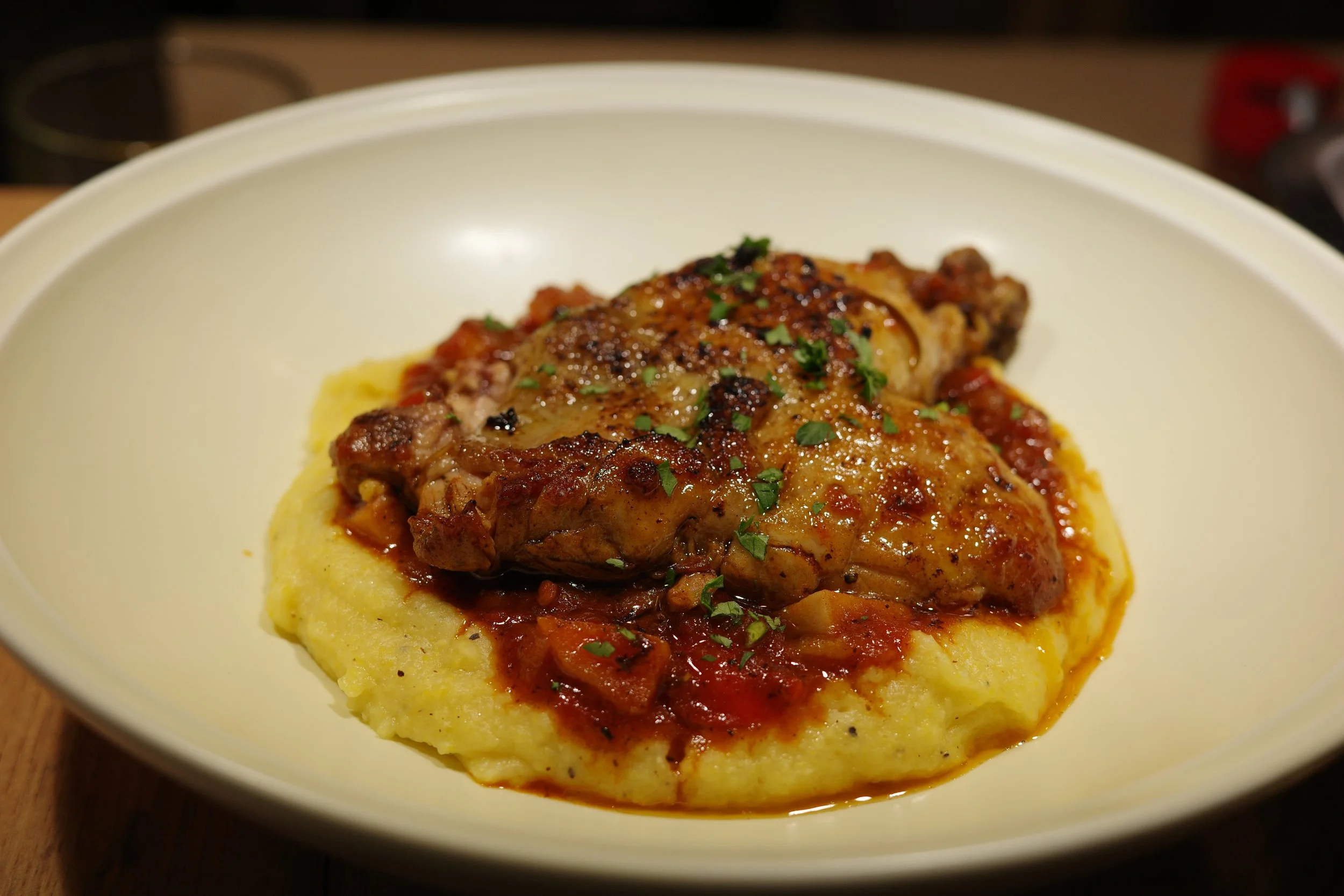 Plate of mashed potatoes topped with braised chicken thigh in tomato sauce, garnished with chopped parsley.