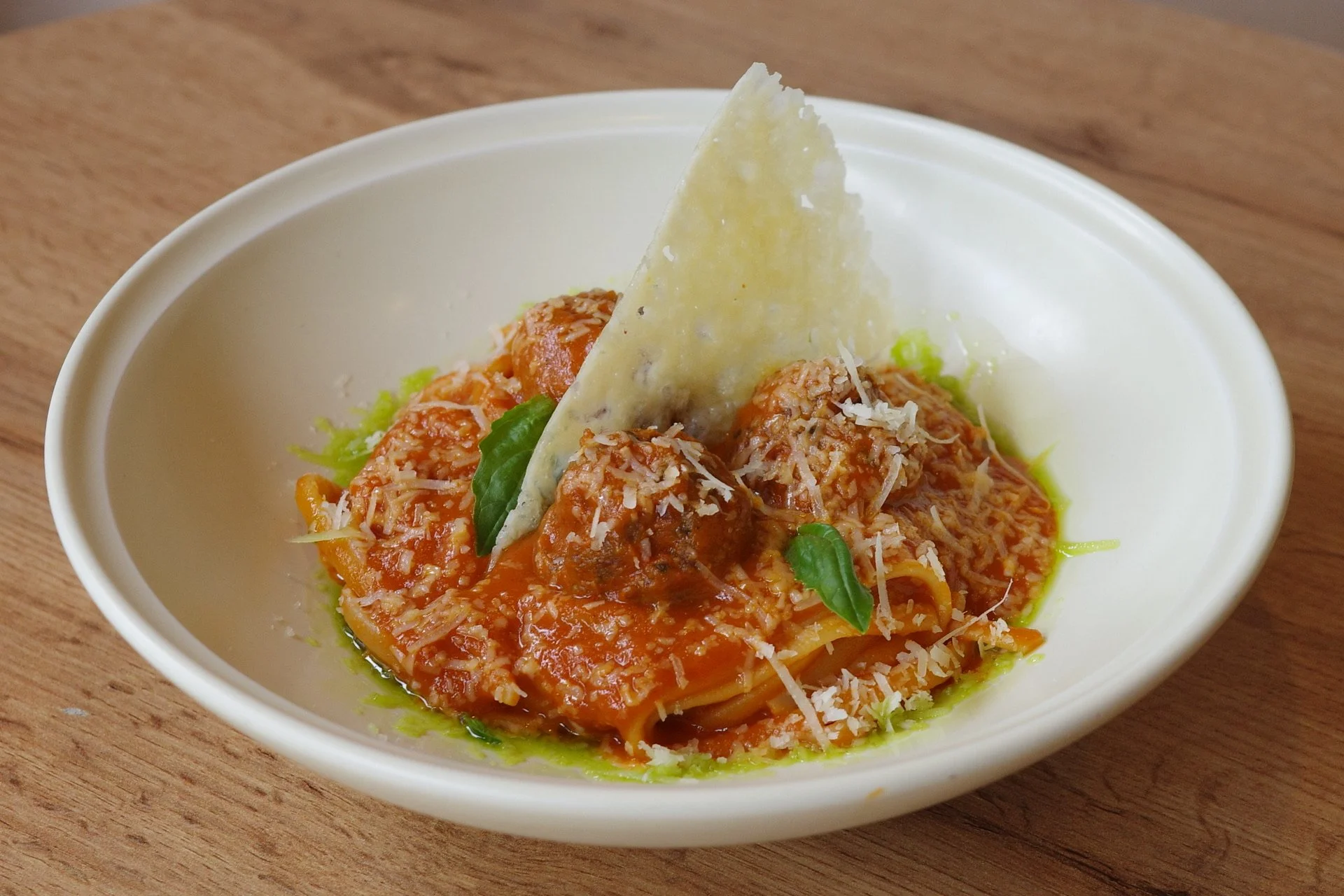 Bowl of spaghetti pasta with meatballs, topped with grated cheese, basil leaves, and parmesan cheese, served in a white bowl on a wooden surface.