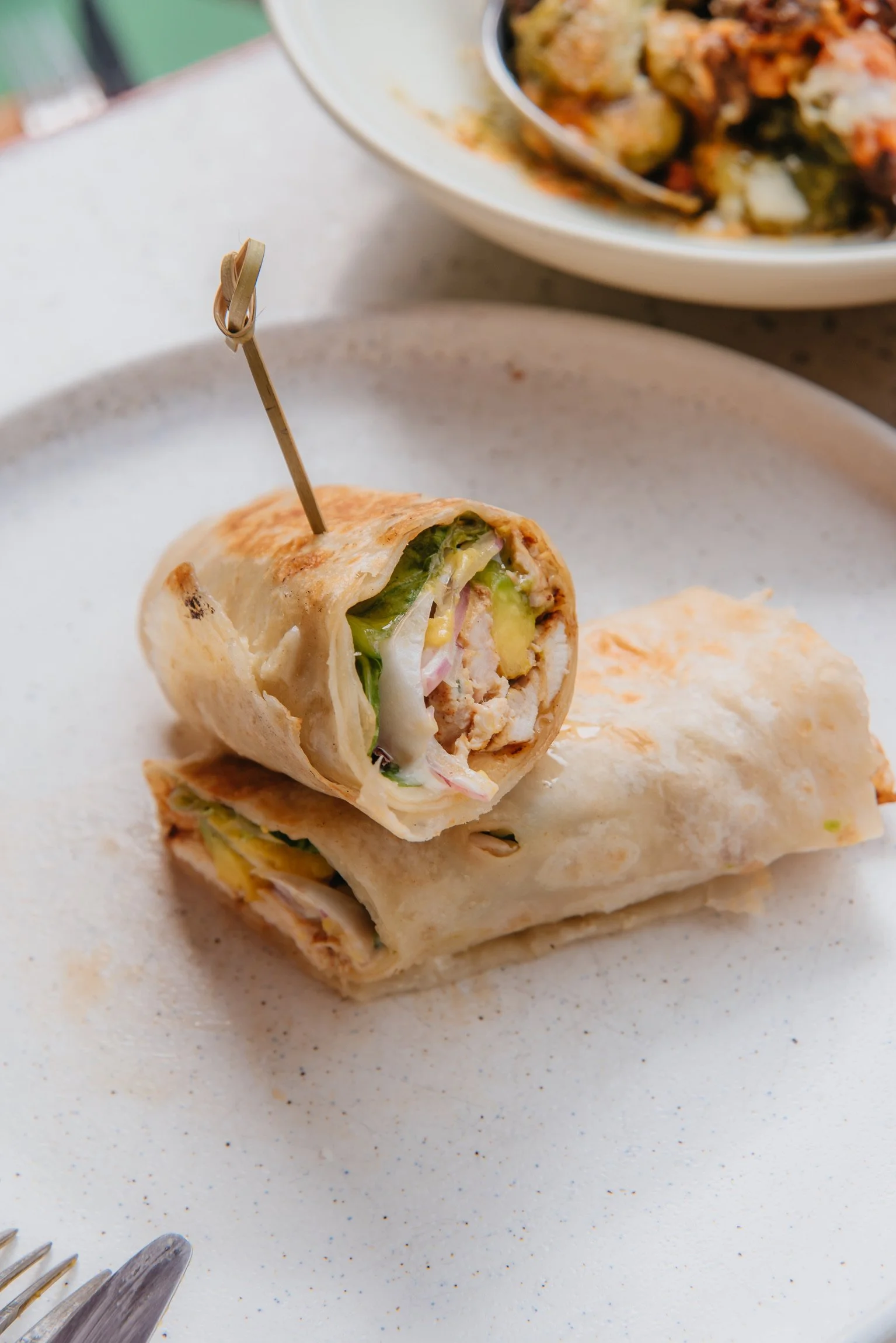 A close-up of a partially eaten burrito on a white plate, with visible ingredients like lettuce, tomato, and meat, and a bowl of additional ingredients or salsa in the background.