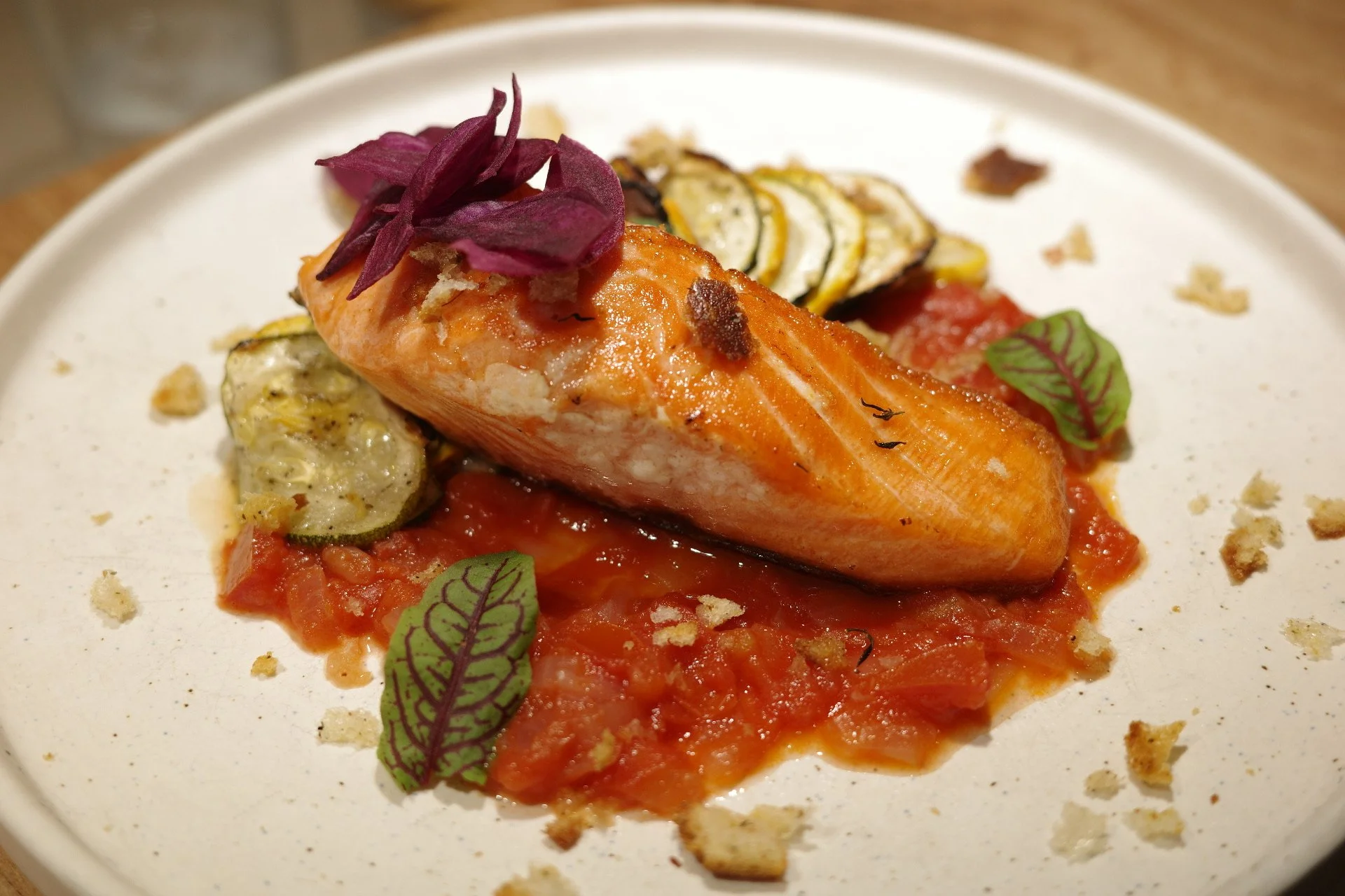 A cooked salmon fillet on a bed of tomato sauce, garnished with thin slices of zucchini, purple pickled onions, and small green leaves, served on a white plate.