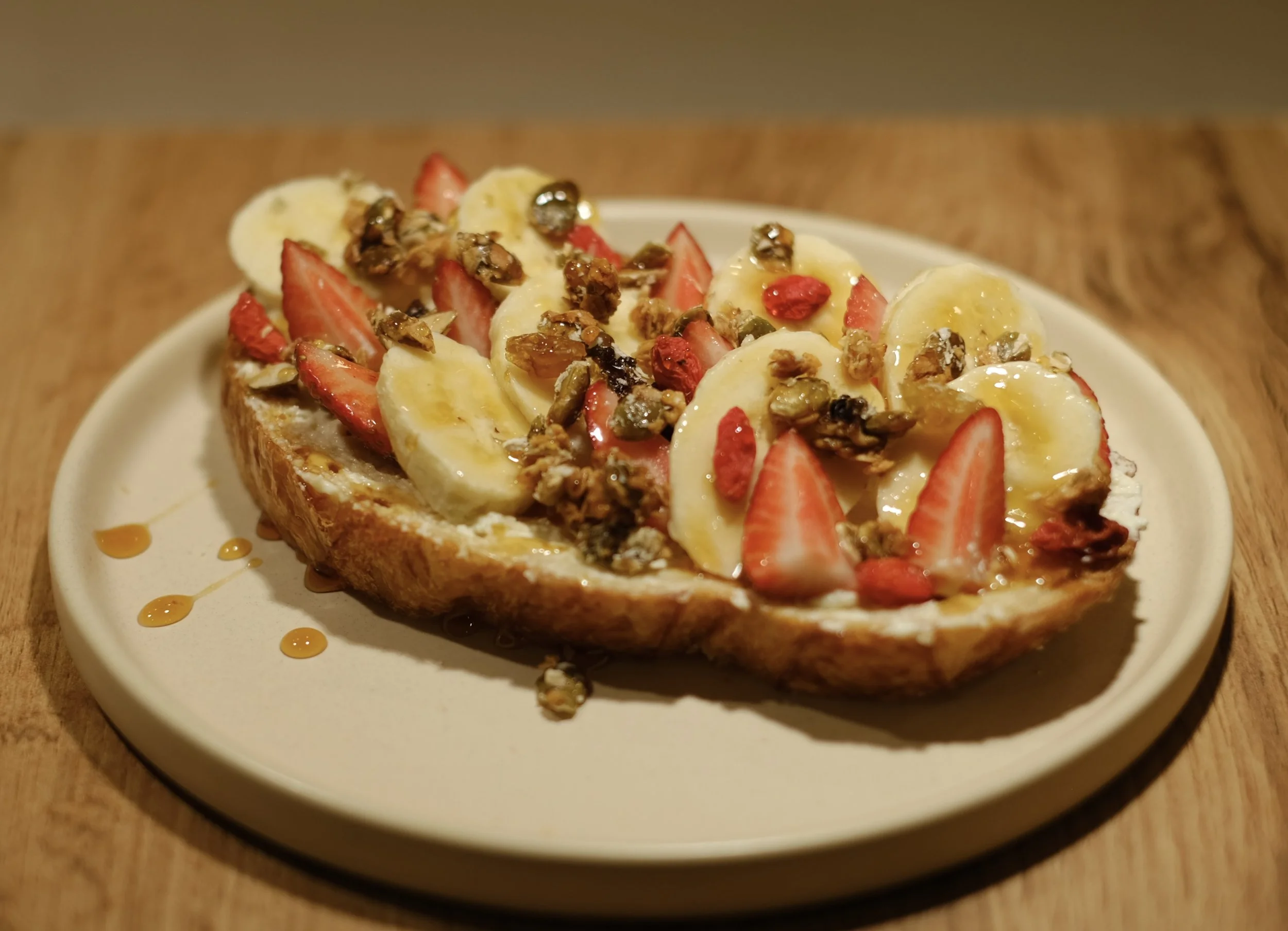 A slice of bread topped with sliced bananas, strawberries, granola, and honey on a white plate.