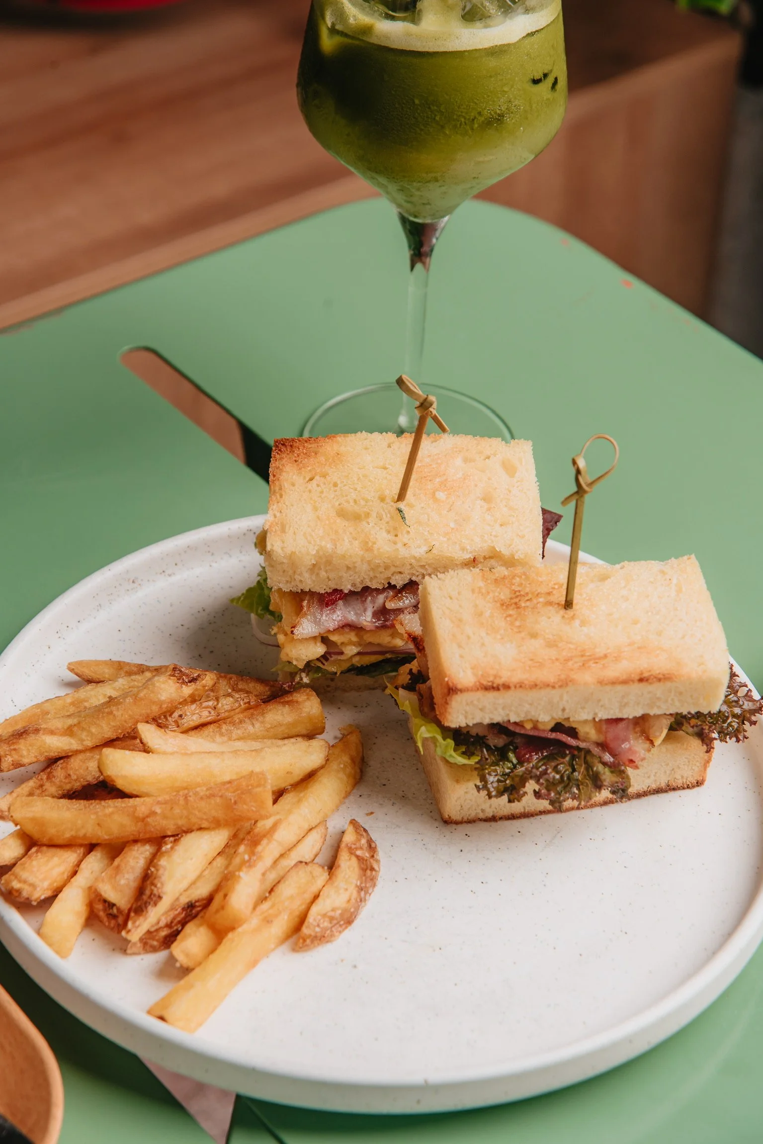 A plate of French fries and a club sandwich on a green table with a green drink in a wine glass.