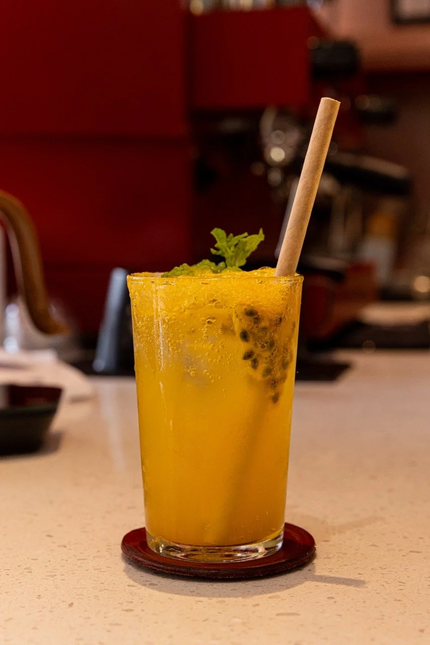 A tall glass of yellow passion fruit cocktail garnished with mint, with a paper straw, on a wooden coaster on a beige countertop.