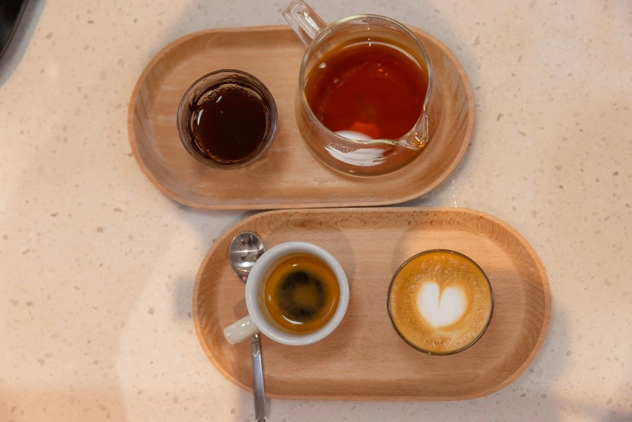 Two wooden trays with various coffee drinks on a speckled white countertop. The top tray has a glass coffee pot and a small glass of black coffee. The bottom tray has a small white cup of espresso with a spoon, and a frothy coffee drink with latte ar