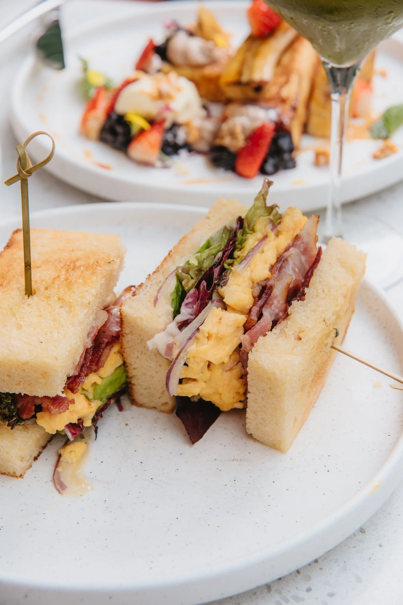 Close-up of bacon, lettuce, tomato, and egg salad sandwich on white bread with a side of toasted bread, served on a white plate.