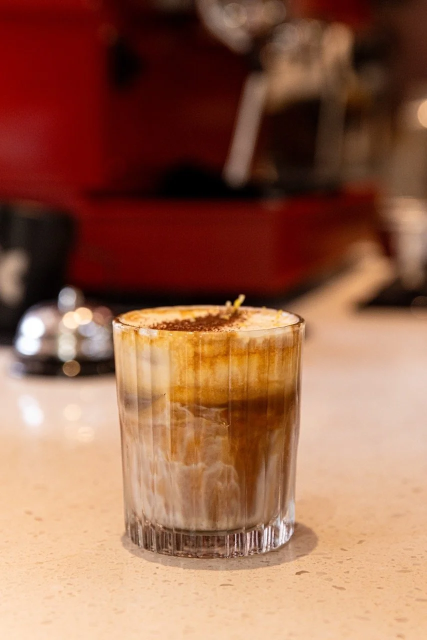 A glass of iced coffee topped with coffee foam and cocoa powder on a counter, with a blurred background.