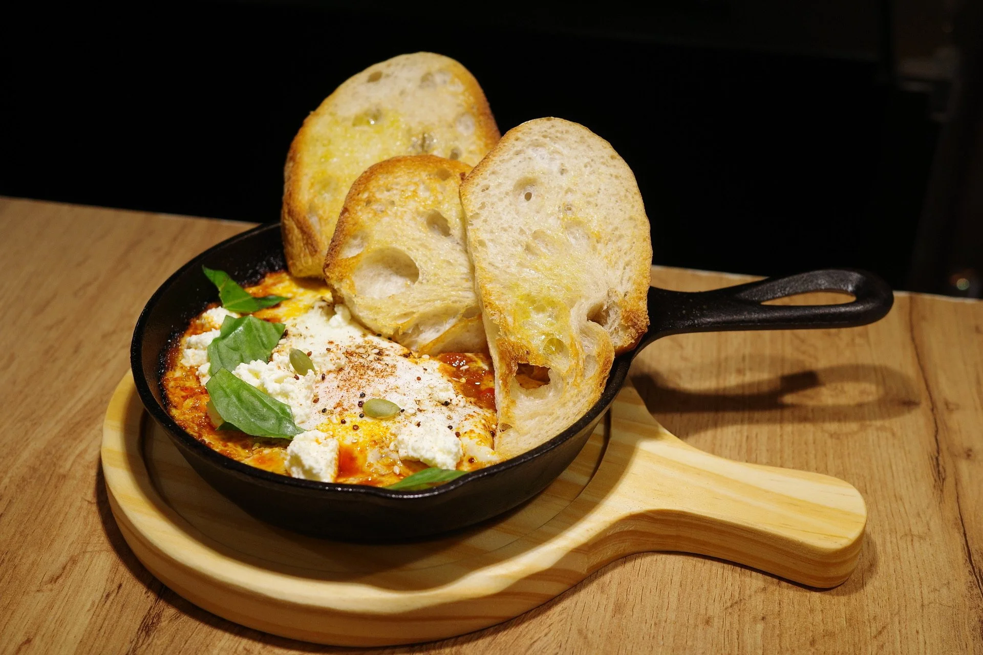 A cast iron skillet of baked eggs with tomato sauce, cheese, basil, and seasoning topped with crispy bread slices on a wooden board.