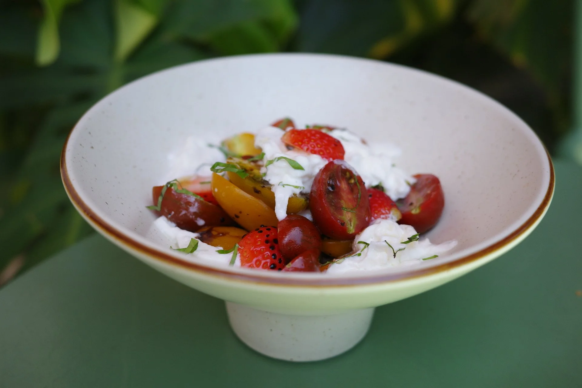 A bowl of fresh strawberries, cherry tomatoes, and a dollop of whipped cream or yogurt, garnished with chopped herbs, placed on a green surface with a background of green leaves.