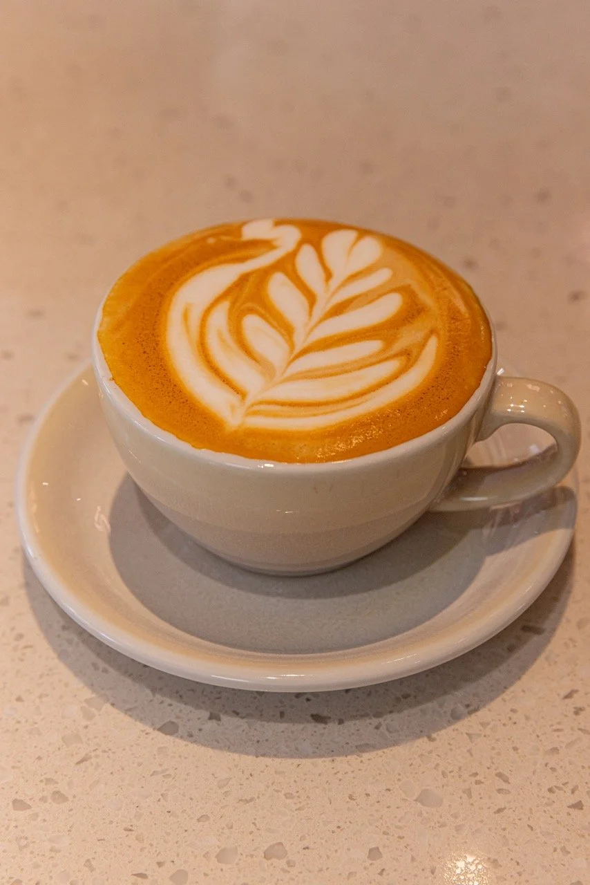 A white cup of coffee with latte art on top, sitting on a matching saucer on a light-colored textured countertop.